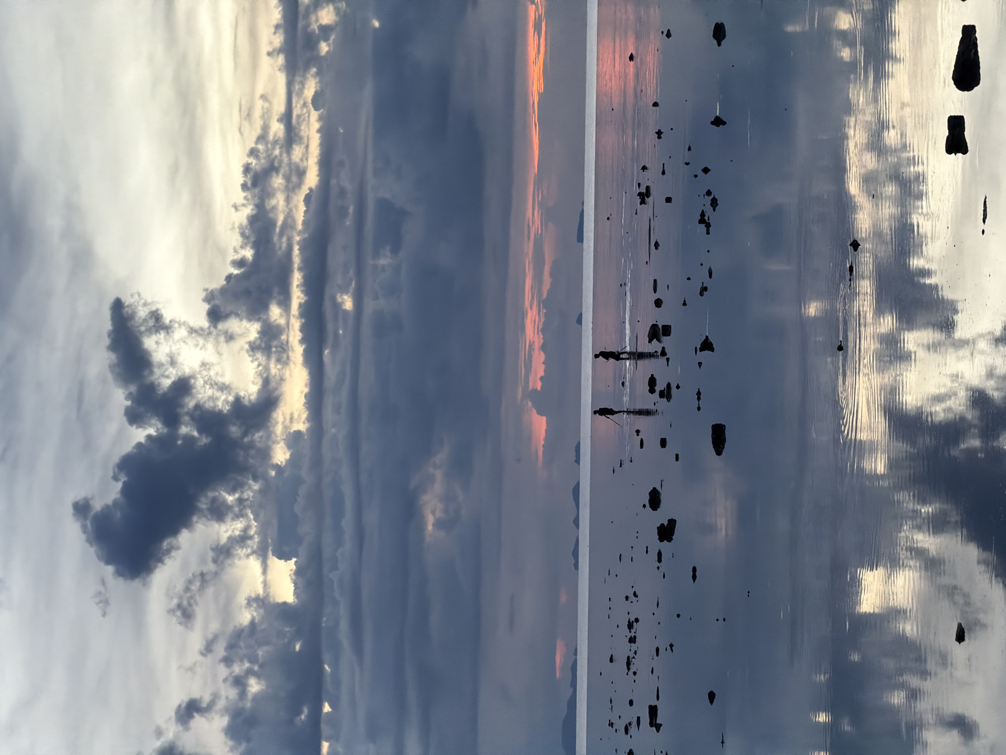 At low tide in Baan Plai Laem, Thailand, two silhouetted figures are wading through shallow water near scattered rocks. They are positioned in the middle ground of the image, with the larger of the two figures slightly to the right. Both figures appear to be holding long poles or spears. The water reflects the overcast sky, which features a dramatic mix of dark gray clouds and patches of soft, warm light, suggesting either sunrise or sunset. In the distance, a faint horizon line separates the water from a series of dark, mountainous landforms. The foreground consists of more rocks partially submerged in the reflective water, creating a mirror-like surface.