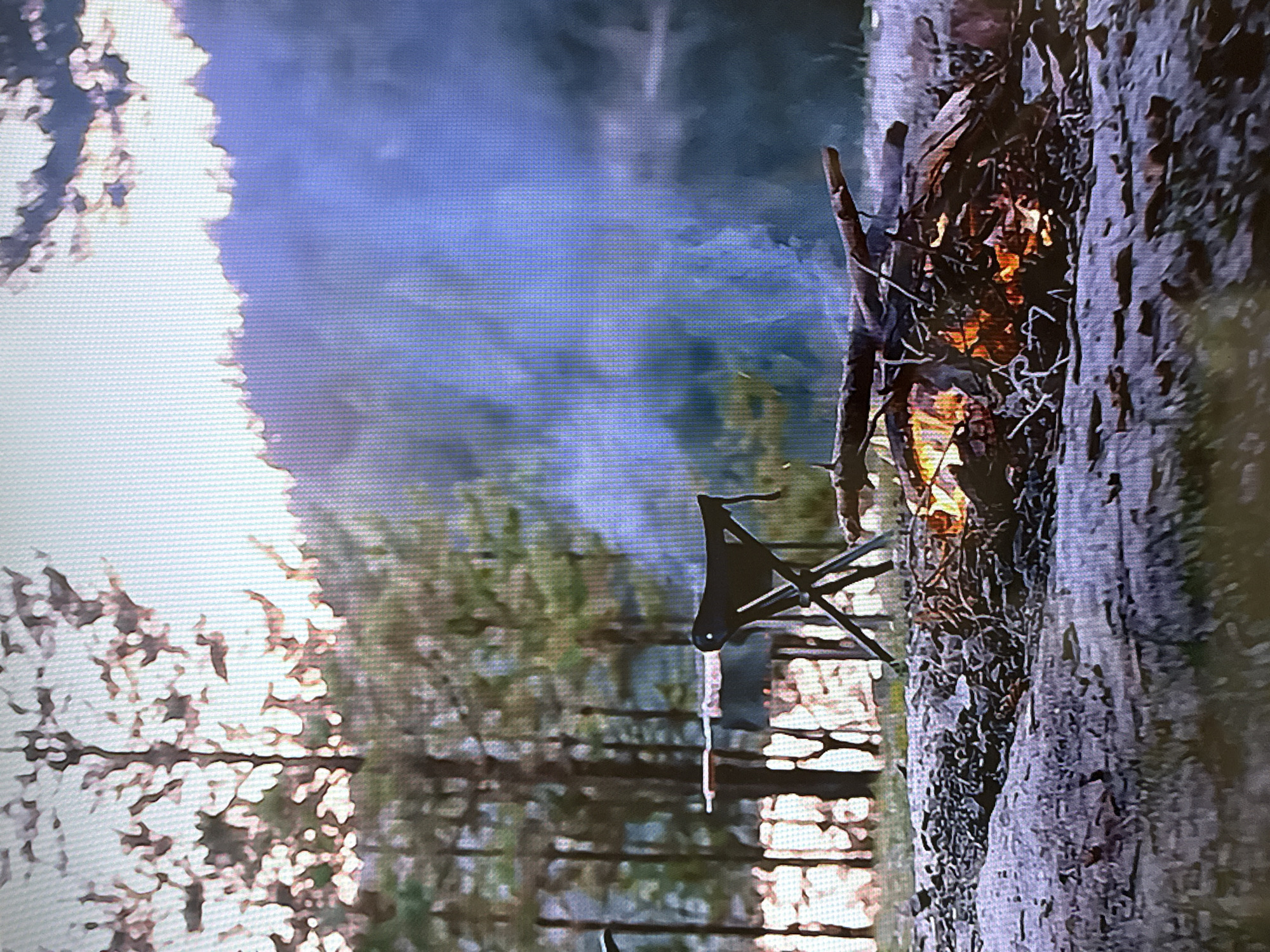 The image depicts an outdoor campsite located in Votkinsk, Russia, during what appears to be dawn or dusk. In the immediate foreground, a smoldering campfire burns with visible orange flames and emits a significant amount of white smoke that drifts towards the background. The fire consists of various logs and smaller branches arranged on bare ground. To the left of the campfire, a black, collapsible three-legged camping stool is set up. Further into the mid-ground, a dark, flat surface, possibly a camping table, is partially visible, positioned amongst sparse vegetation. The background features a dense forest of evergreen trees, many appearing as dark silhouettes against a bright, hazy sky. Beyond the trees and obscured by smoke and atmospheric haze, a large body of water, likely a lake or wide river, is discernible. No individuals are present in the scene. The overall environment suggests a tranquil natural setting prepared for or actively hosting outdoor recreation.