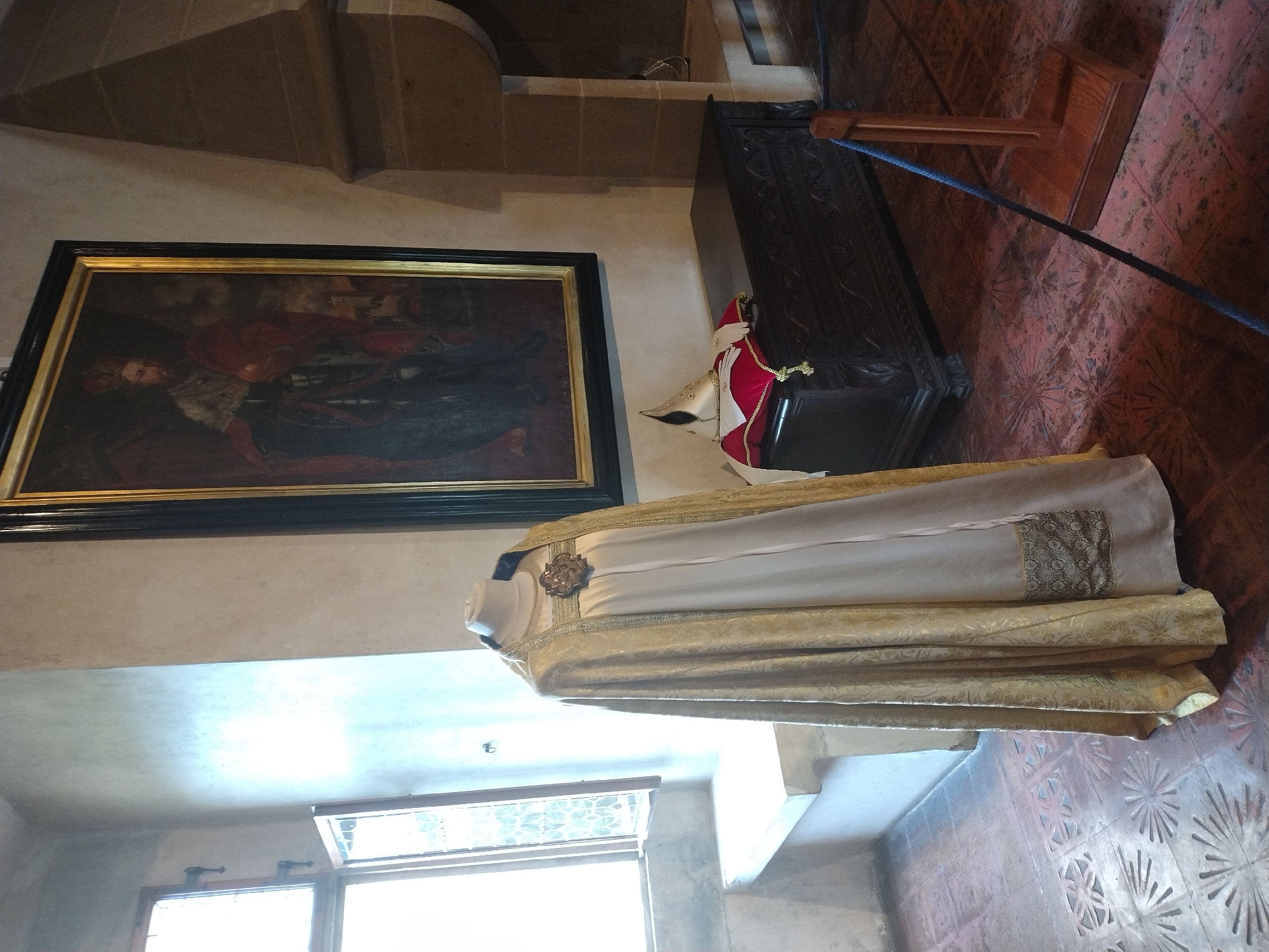 The image shows a room in Karlštejn Castle, Czechia, likely a museum exhibit. In the foreground, a mannequin is dressed in an ornate, light-colored robe with a gold patterned cape draped over it. A decorative brooch is fastened at the neck. Behind the mannequin is a dark wooden chest with intricate carvings, topped with a white and gold ceremonial hat resting on a red cushion. A large, framed painting of a regal figure in red and gold attire hangs on the wall behind the chest. To the left, a window with leaded glass panes allows natural light to enter the room, illuminating a stone bench beneath it. The floor is covered with red tiles featuring a decorative pattern. A rope barrier on a wooden stand is visible in the lower right corner of the image. The overall atmosphere suggests a historical display.