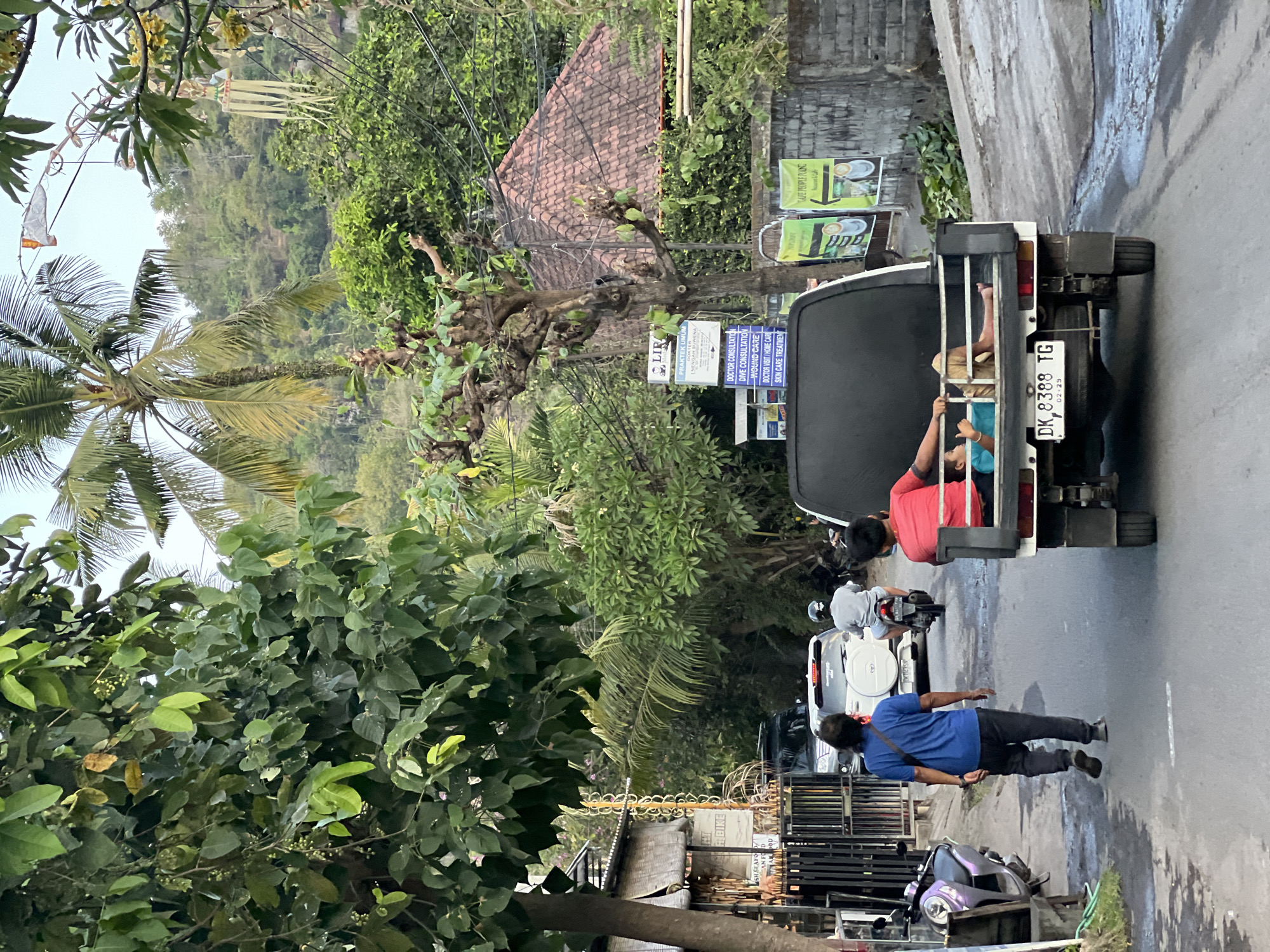 In Abang, Indonesia, a narrow paved road runs between lush green trees and buildings. A man in a blue shirt and black pants walks away from the camera down the road. Behind him, a white SUV and a motorcycle carrying one rider are visible. In the foreground, a white pickup truck with a black canopy is moving away from the camera. A child in a teal shirt and tan shorts lies in the back of the pickup truck, resting their head on their arm. The child appears relaxed. The overall scene depicts a typical day on a street in a tropical locale, with vehicles and pedestrians navigating the roadway amidst abundant vegetation.