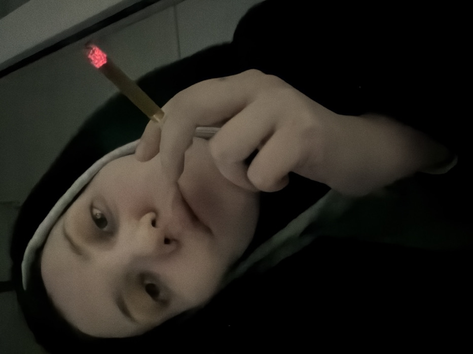A selfie shows a person reclining, wearing a black hooded sweatshirt with a white drawstring. They are holding a lit cigarette or joint in their right hand, the tip glowing red. The person is fair-skinned, with dark eyes and a neutral expression. The background is dark and indistinct, suggesting an indoor setting with limited light. The overall image has a casual, informal quality.