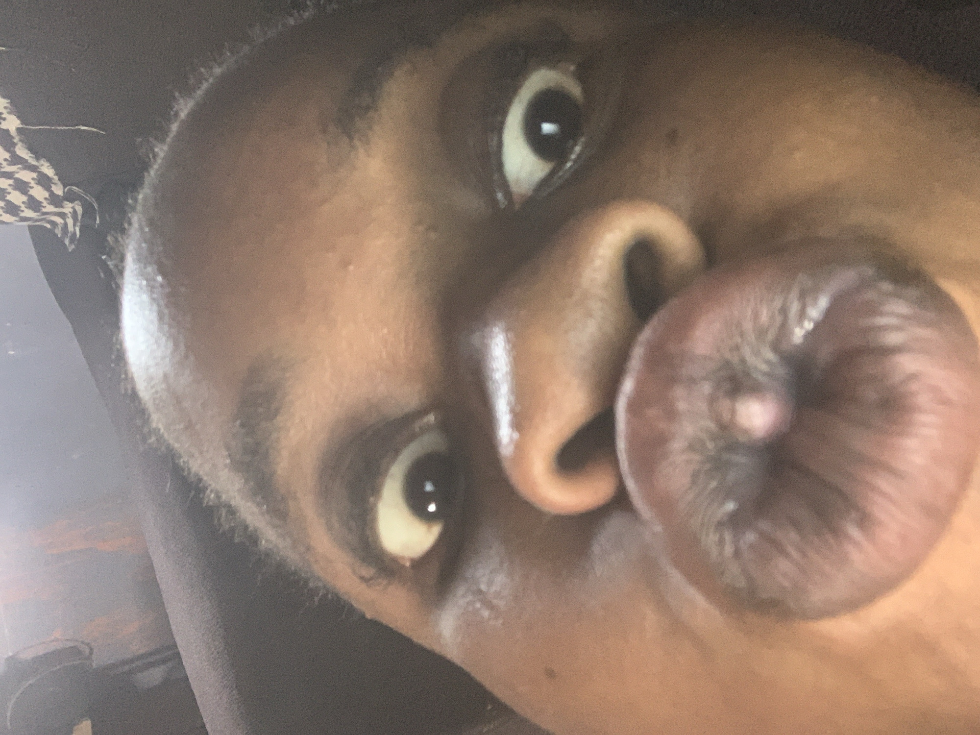 A close-up, angled selfie shows a person of color with their lips pursed into a round pout. The person's eyes are wide and looking directly at the camera. The background is out of focus, showing glimpses of clothing.