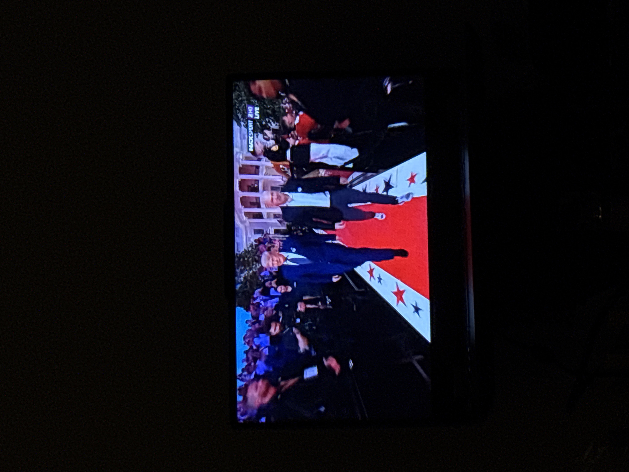 The image shows a television screen displaying a scene from an event. Two men are walking on a red carpet decorated with white and red stars. The man on the left is wearing a blue suit and tie, while the man on the right is wearing a dark blazer over a light-colored shirt and dark pants. There are other people in the background, some of whom appear to be security personnel. A building resembling the White House is visible in the background. The text "MAXSPORT 2HD LIVE" is visible in the upper right corner of the screen. The image is taken in the city of Enschede, Netherlands.