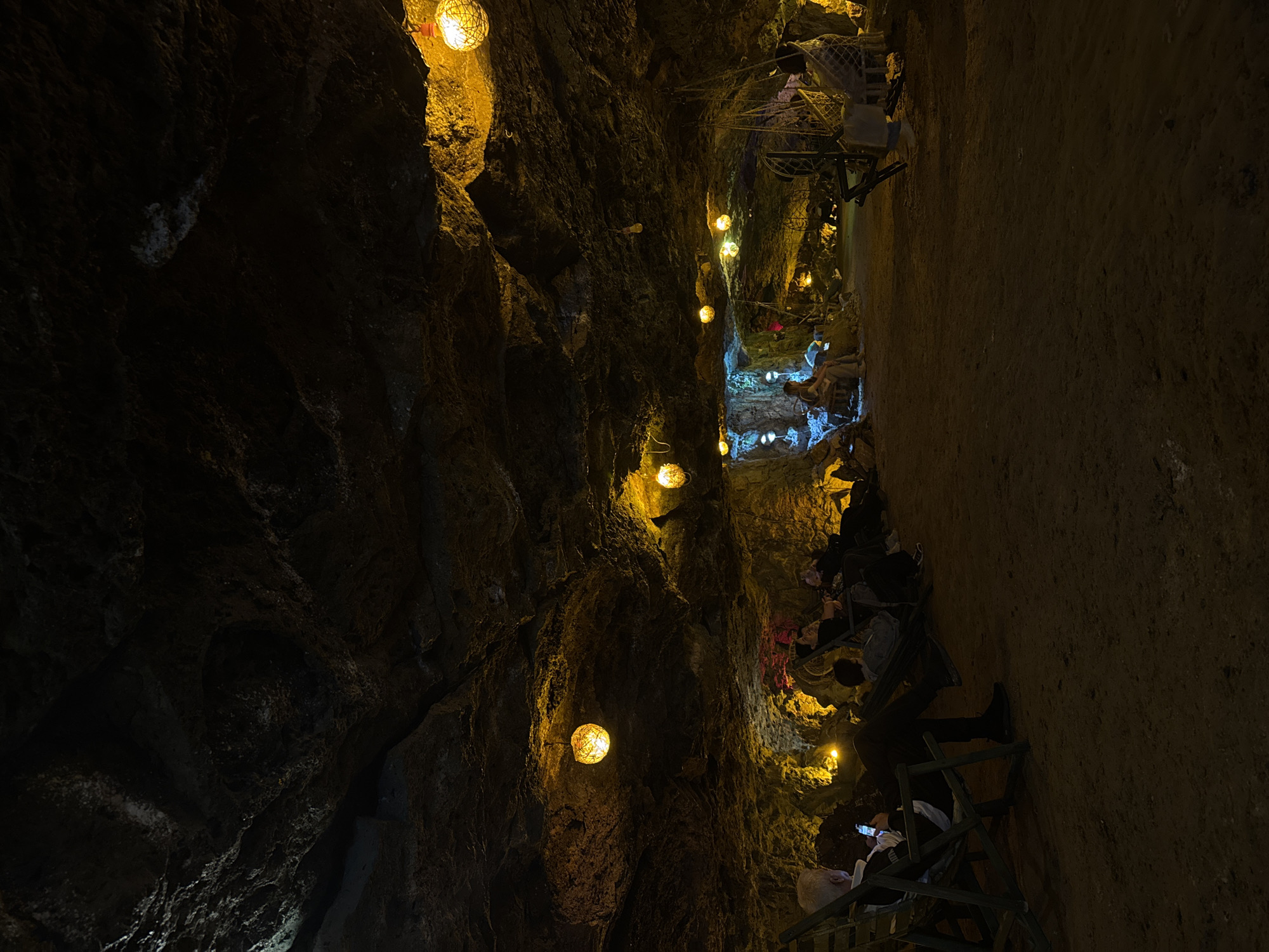 The image depicts a group of people inside a dimly lit cave or underground space, likely a tourist attraction in Alaverdi, Armenia. The cave walls are rough and textured, with natural rock formations visible. The scene is illuminated by numerous small, spherical lights with a warm, yellow glow, hanging at various heights and along the path. Some of these lights appear to be encased in a mesh or woven material.
The people are seated on what look like tiered benches or steps, positioned along the cave. Many are looking forward or down, some are holding phones, suggesting they are observing something or participating in an experience. The atmosphere appears relaxed and contemplative, with the soft lighting creating a cozy and somewhat mysterious ambiance.
The pathway through the cave is narrow and winding, carved out of the rock. The ground appears to be dirt or gravel. The overall impression is that of a curated underground space designed for visitors to explore and experience. There are no clear indications of the time of day or weather, as the setting is entirely enclosed. No visible text is present in the image.