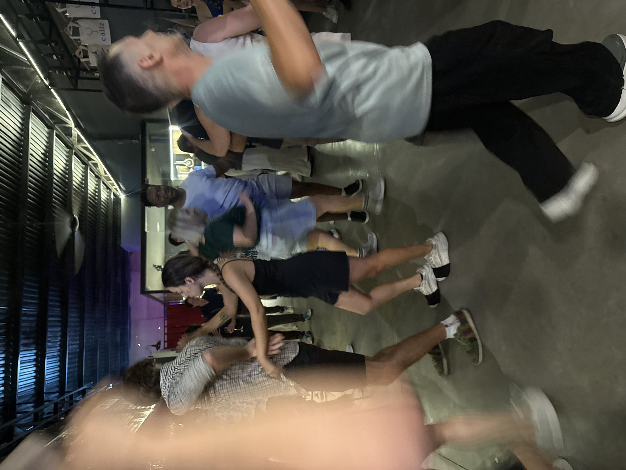 The image shows a group of people participating in a physical activity, likely a dance or group exercise. They are arranged in a line, with each person holding onto the shoulders of the person in front of them. The people are of various ages and ethnicities. The location appears to be an indoor venue with exposed rafters and dim lighting. There are signs on the wall.