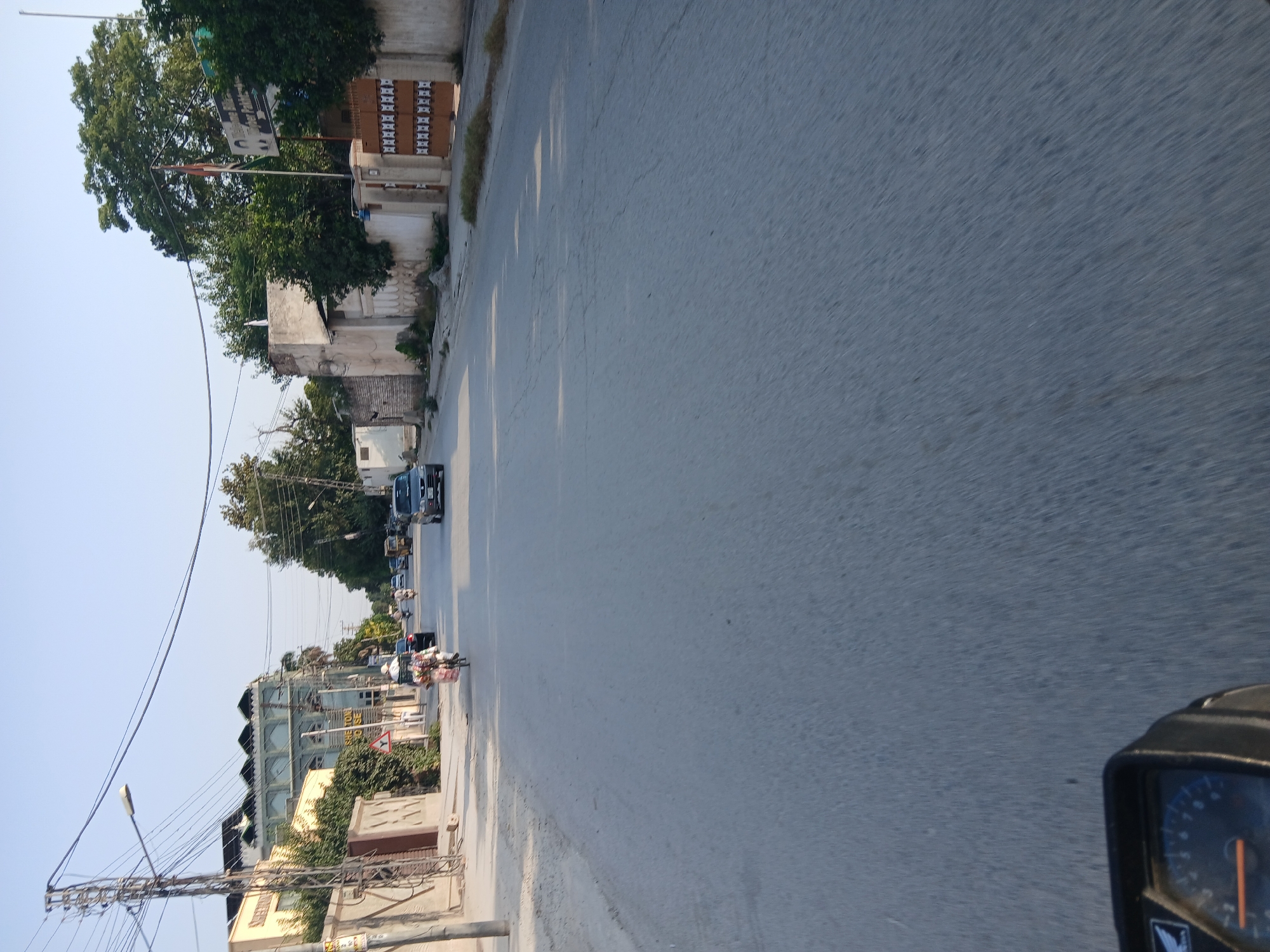 The image shows a street scene in Peshawar City Tehsil, Pakistan. In the foreground, a portion of a motorcycle's dashboard is visible. The road itself is paved and appears to be in moderate condition with some cracks. On the left side of the road, there is a utility pole with multiple wires and a street lamp. Adjacent to the pole is a building with a light-colored facade and windows. Further down the street on the left, a more prominent building with a distinctive architectural style, featuring arched windows and a dark green roof, is visible. A smaller sign on this building reads "SHEHTON HOUSE".
On the right side of the road, there is a wall with decorative elements and a large brown gate. Behind the wall, trees and other buildings are present. There are several vehicles on the road, including a car, a motorcycle with a person and a load of goods, and possibly other vehicles further in the distance. The sky is clear and light blue, suggesting it is daytime. The overall impression is a typical urban street in Pakistan.