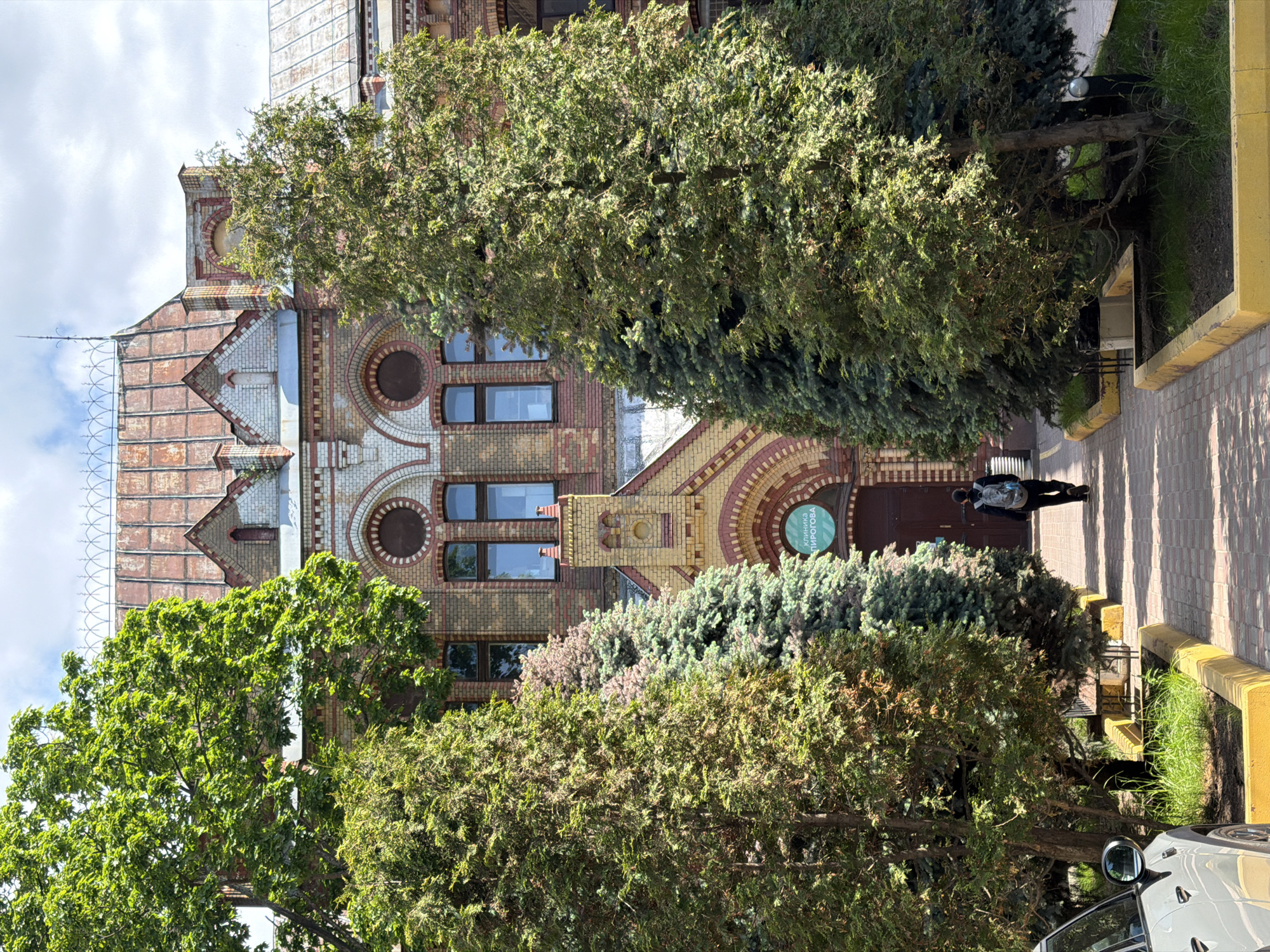 This upright image captures a detailed brick building partially hidden by lush green trees and bushes, under a bright, cloudy daytime sky. The multi-story building exhibits intricate architectural designs, featuring decorative brick patterns in hues of red and tan, including arched windows and prominent circular motifs. Its roof presents a distinctive gabled structure with patterned shingles, and a slender antenna-like fixture rises from the left side of the roofline.
In the foreground, a paved sidewalk runs alongside a vibrant yellow curb and a narrow strip of greenery. A sole individual, dressed in dark attire and carrying a light-colored backpack, is walking away from the building's entrance on the sidewalk. The entrance itself is a dark wooden door set within an archway. Adjacent to the entrance, a circular sign is mounted on the building, displaying the Cyrillic text "КЛИНИКА ПИРОГОВА," which translates to "Pirogov Clinic."
Numerous trees with dense green foliage dominate the foreground and midground, casting shadows on the pedestrian area and partially obscuring the building's lower and mid sections. Both deciduous and evergreen trees are present. A portion of a white car is visible in the bottom right corner, peeking from behind the curb and vegetation. The soft, diffused lighting indicates it is daytime, possibly late morning or early afternoon, with an overcast sky. The scene depicts an urban environment, combining historical architecture with significant natural elements.
