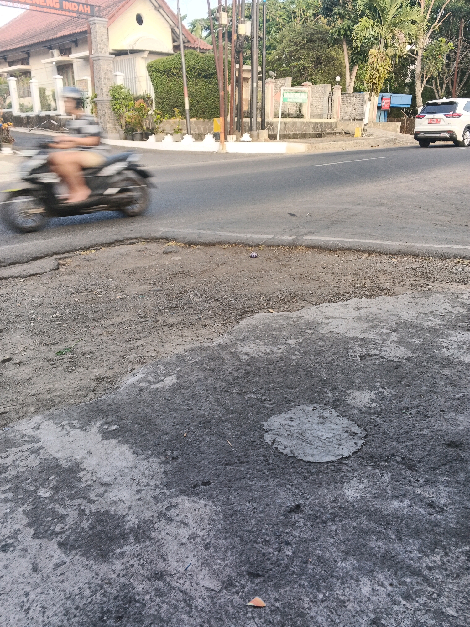 A person is riding a motorcycle on a street in Semarang, Indonesia. The motorcycle is moving from left to right and is blurred due to motion. The rider is wearing a helmet and shorts. In the background, there are buildings, trees, and a white car. The road appears to be in disrepair with patches of asphalt missing.
