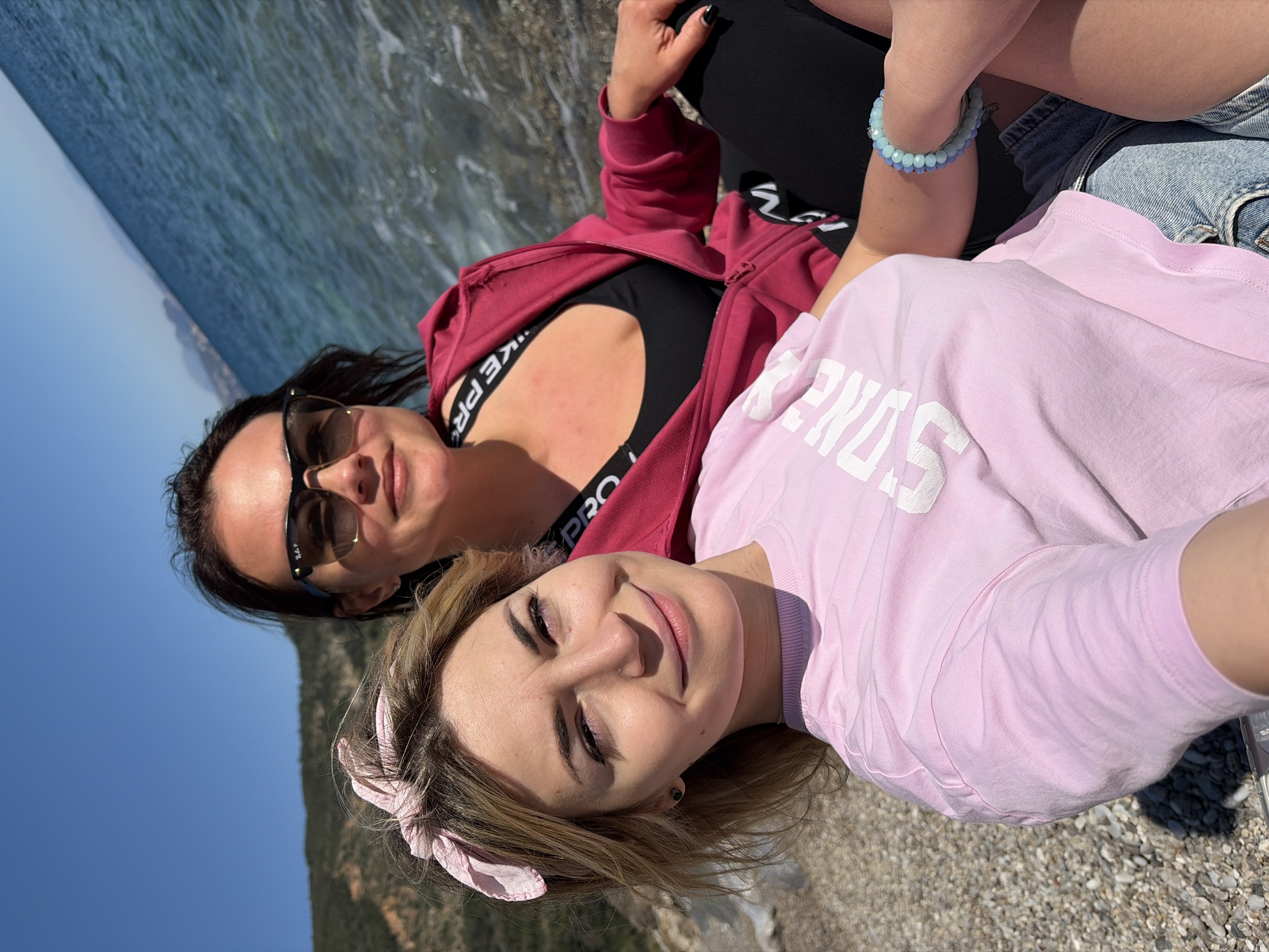 Two women are reclining on a pebble-covered beach in Prijevor, Montenegro. The woman in the foreground wears a light pink t-shirt with white lettering, a pink ribbon in her hair, and has light brown hair. The woman in the background is wearing sunglasses, a red jacket, and a black sports bra. They appear to be near the shoreline with blue water visible. The sky is blue with some land in the distance.