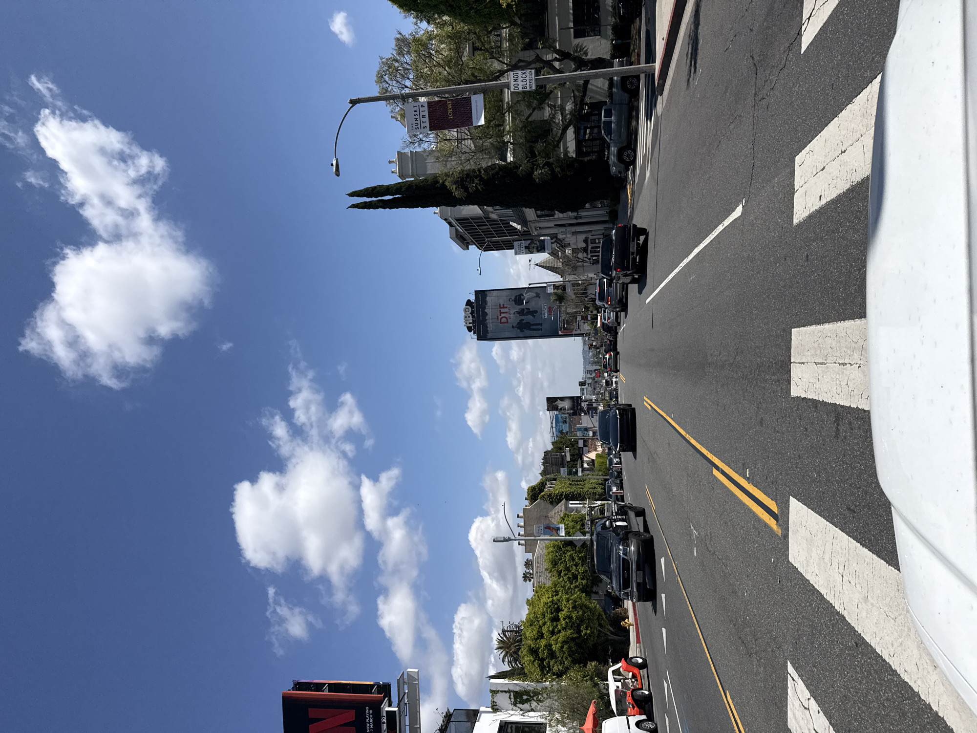The image depicts a street scene in Los Angeles, California. A two-lane road with double yellow lines running down the center is visible, along with crosswalk markings. There's moderate traffic consisting of cars and a small recreational vehicle. On the left side of the road, there are buildings with billboards for Netflix and HBO. Street signs and light posts are also present. In the background, more buildings and trees are visible under a clear blue sky with scattered clouds. A "Sunset Strip" banner is also seen attached to one of the streetlights. The photo is taken from the perspective of a car stopped at a crosswalk.