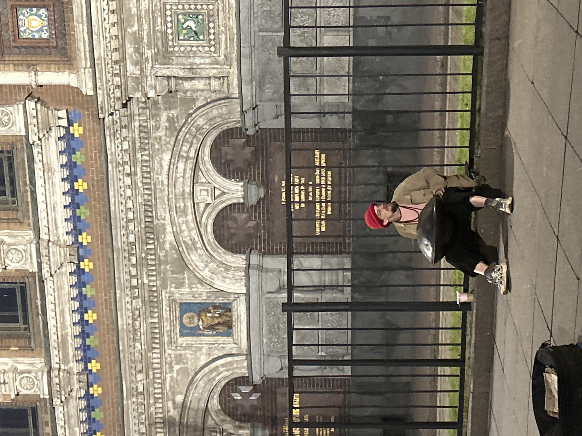 In the city of Unknown, Unknown, a man wearing a red beanie, a tan jacket, and dark pants is sitting on a curb in front of a black metal fence. He is playing a handpan, a percussion instrument. To his left, on the sidewalk, is an open black bag with some papers inside, and a pink cup. The background features a large, ornate building with intricate architectural details, including arched windows, decorative friezes, and colorful tilework. The lighting suggests it is evening or night.
