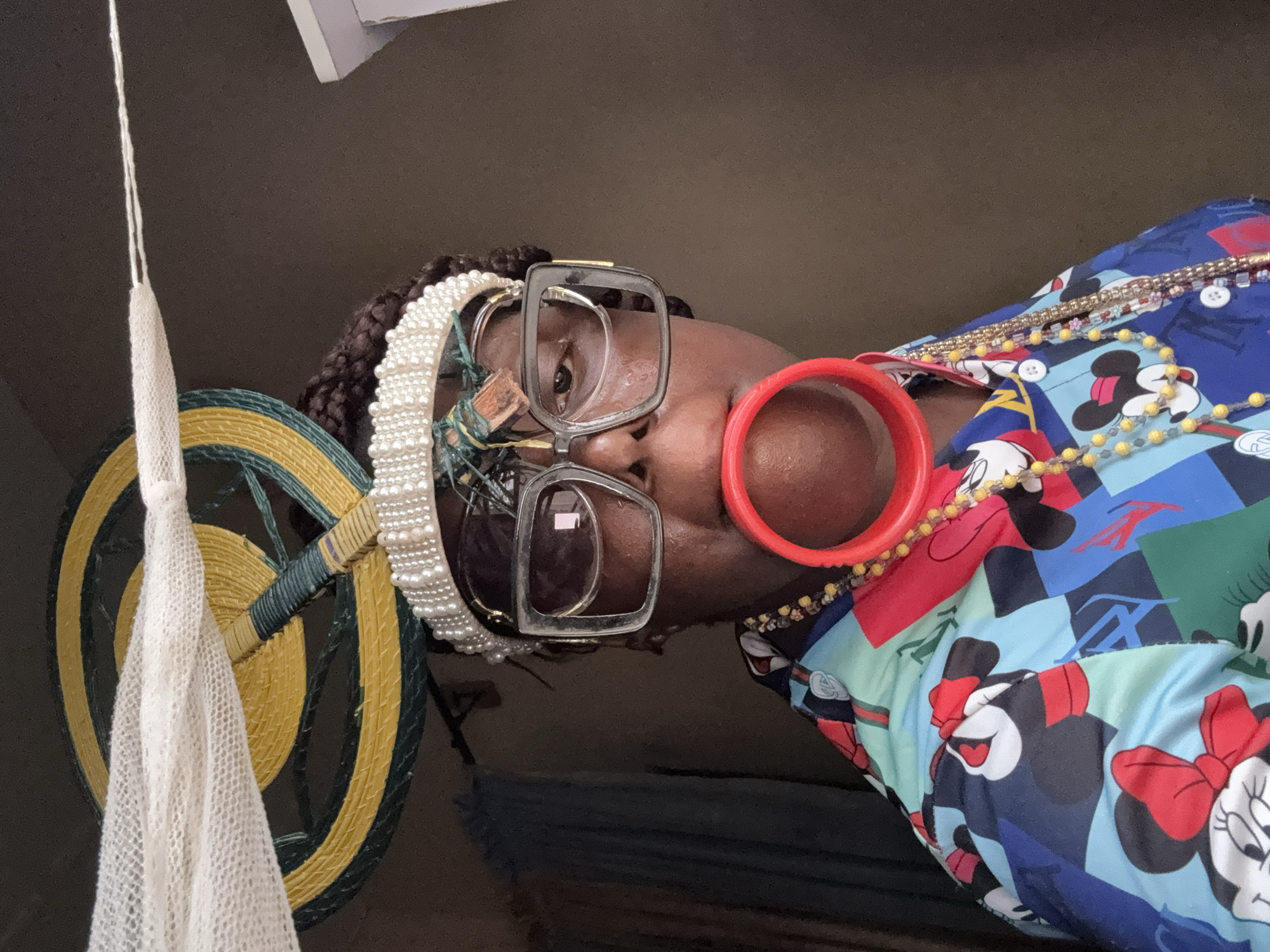 A woman wearing oversized, rectangular eyeglasses, a pearl headband, and a large red ring-shaped earring poses indoors. She is wearing a multi-colored shirt printed with Mickey and Minnie Mouse patterns, adorned with strands of beads and a jeweled necklace. To the left is a decorative, circular wall hanging made of woven material, featuring yellow and green concentric circles and a draping white crocheted tassel. The background is dimly lit and appears to be a plain interior wall. No evidence suggests that the location is Madangala, Nigeria.