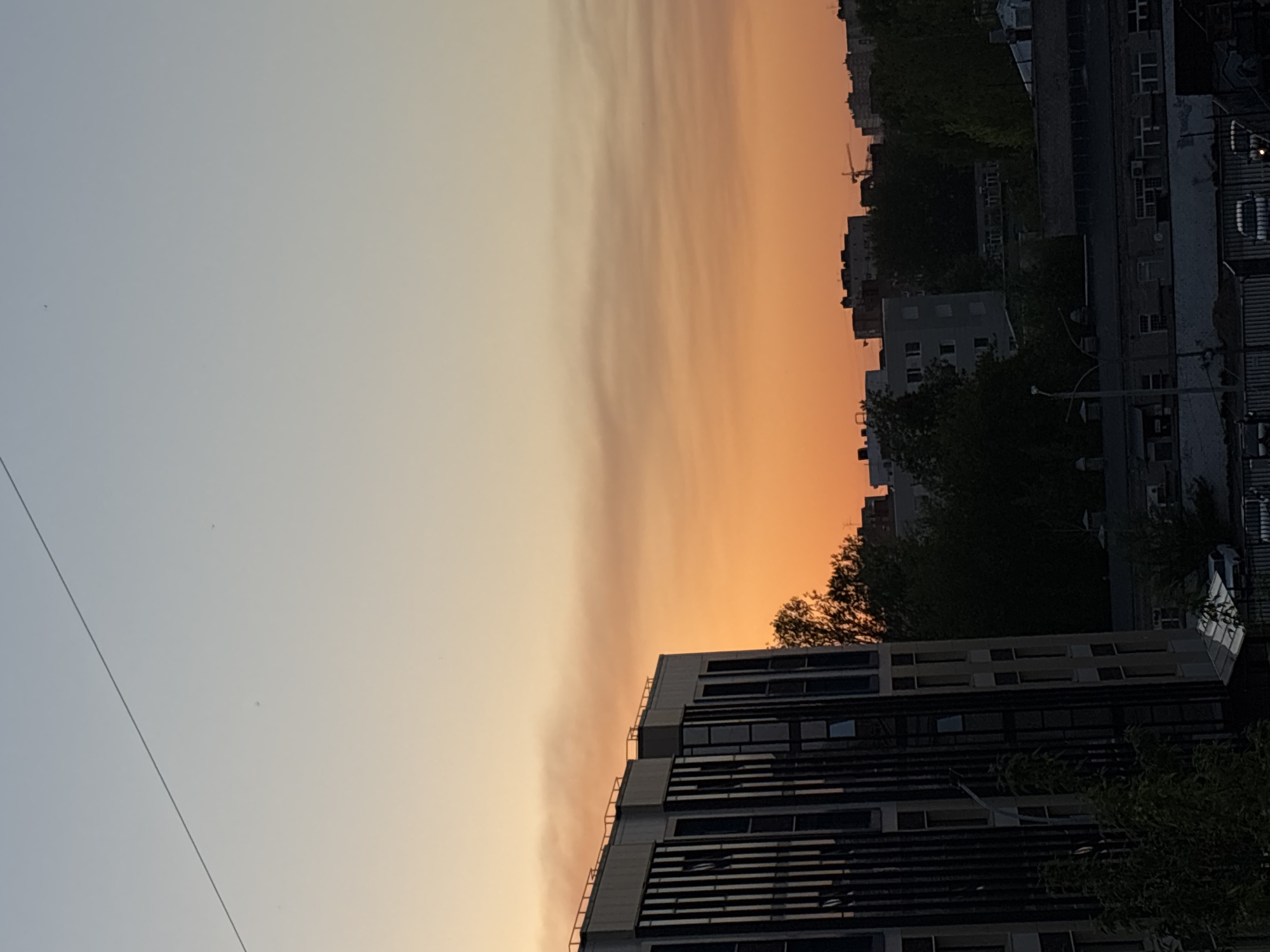 The image depicts a sunset in what appears to be a densely populated urban area, possibly Perm, Russia. The sky transitions from a pale blue in the upper left to a vibrant orange and yellow towards the center and right, indicative of the setting sun. Silhouettes of buildings and trees are visible against the colorful sky. The architecture is predominantly modern, with multi-story buildings and a high-rise structure, indicating that these are a place of urbanity. The image has a strong sense of depth and scale, emphasizing the size of the buildings and the vastness of the sky. There is no evidence of human presence or activity in the immediate foreground.