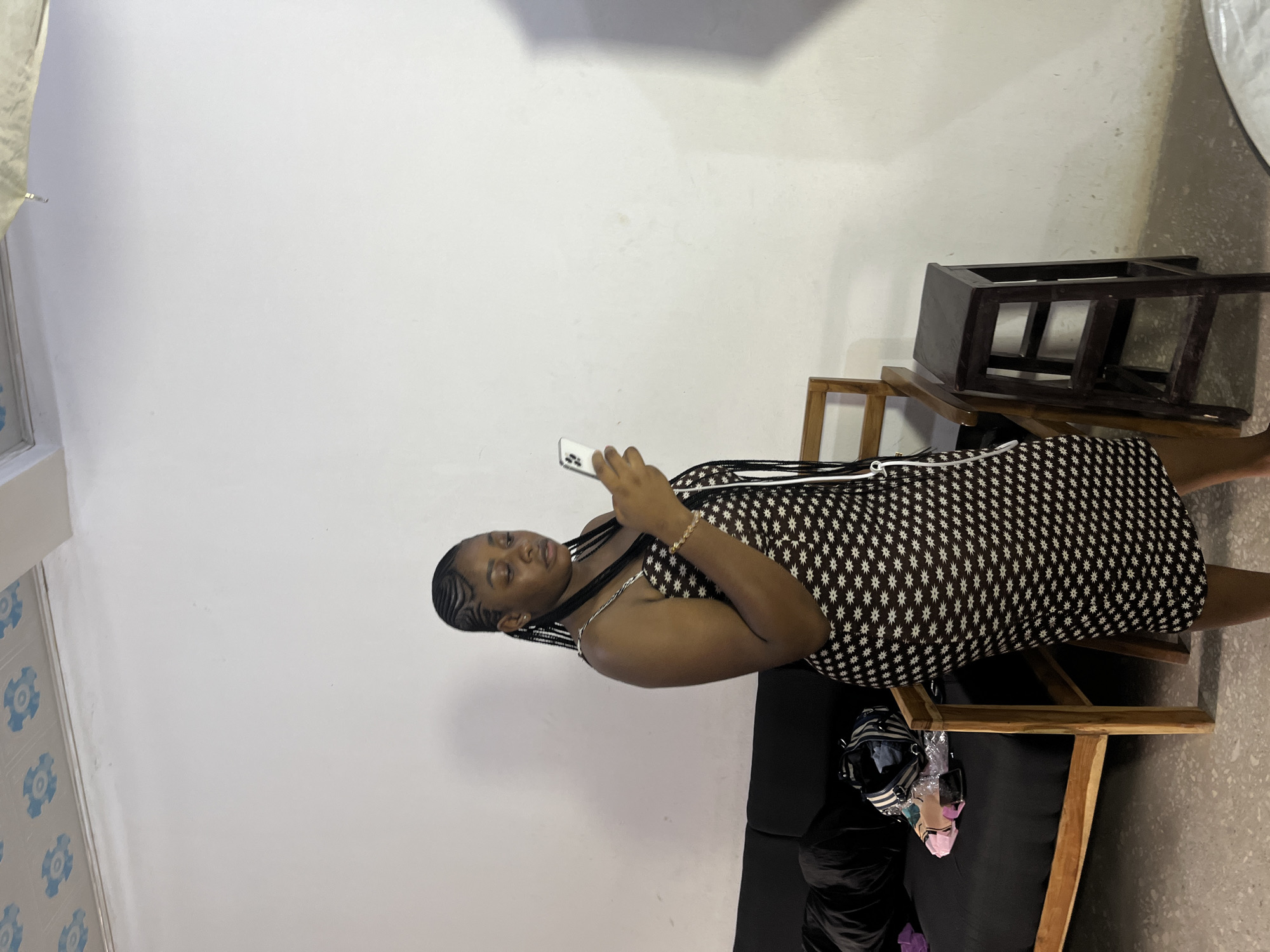 A woman with braids and a patterned dress is looking at her phone. She is sitting on a black couch, next to a wooden chair. There are other chairs and a white wall in the background. The lighting suggests it is daytime.