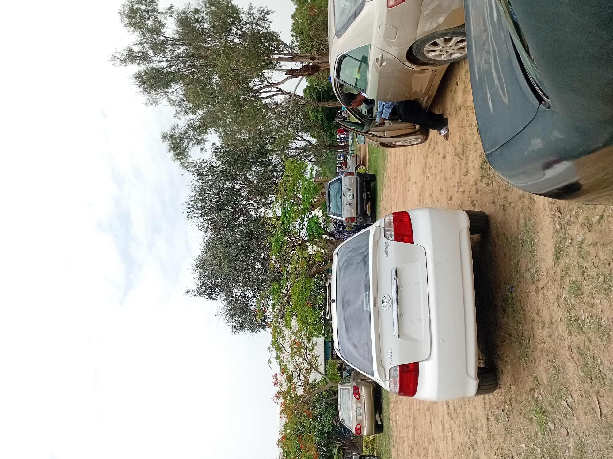 The image depicts a parking area in Jos, Nigeria. Several cars are parked on a dirt and grassy surface under trees. A white Toyota Yaris is in the foreground, with other vehicles of different colors and models visible in the background. A person is exiting a tan-colored car, with the car door open. The sky is overcast.