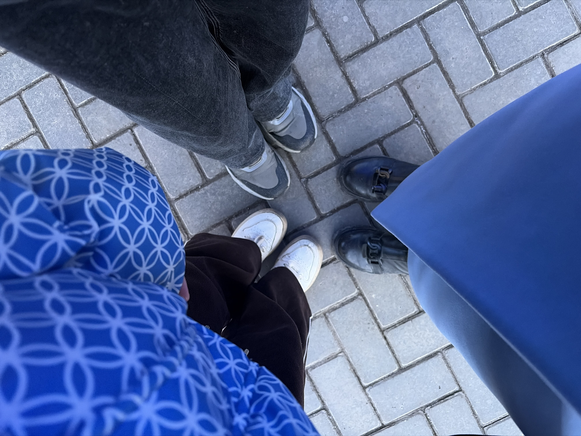 Three individuals are depicted in a top-down view, standing on an outdoor paved surface in Izhevsk, Russia. The ground consists of light grey rectangular paving stones with visible grout lines.
One individual, positioned in the upper-left, wears a blue garment with a repetitive white geometric pattern, brown lower trousers, and white low-top sneakers. A second individual, on the right, wears dark grey denim trousers and grey athletic shoes with white soles. The third individual, at the bottom-center, wears a solid blue garment or accessory and black formal shoes with a metallic buckle detail on each. All three are stationary, with their feet clustered in the frame, suggesting they are standing in close proximity and possibly conversing. No other objects or specific environmental features are discernible beyond the paving.