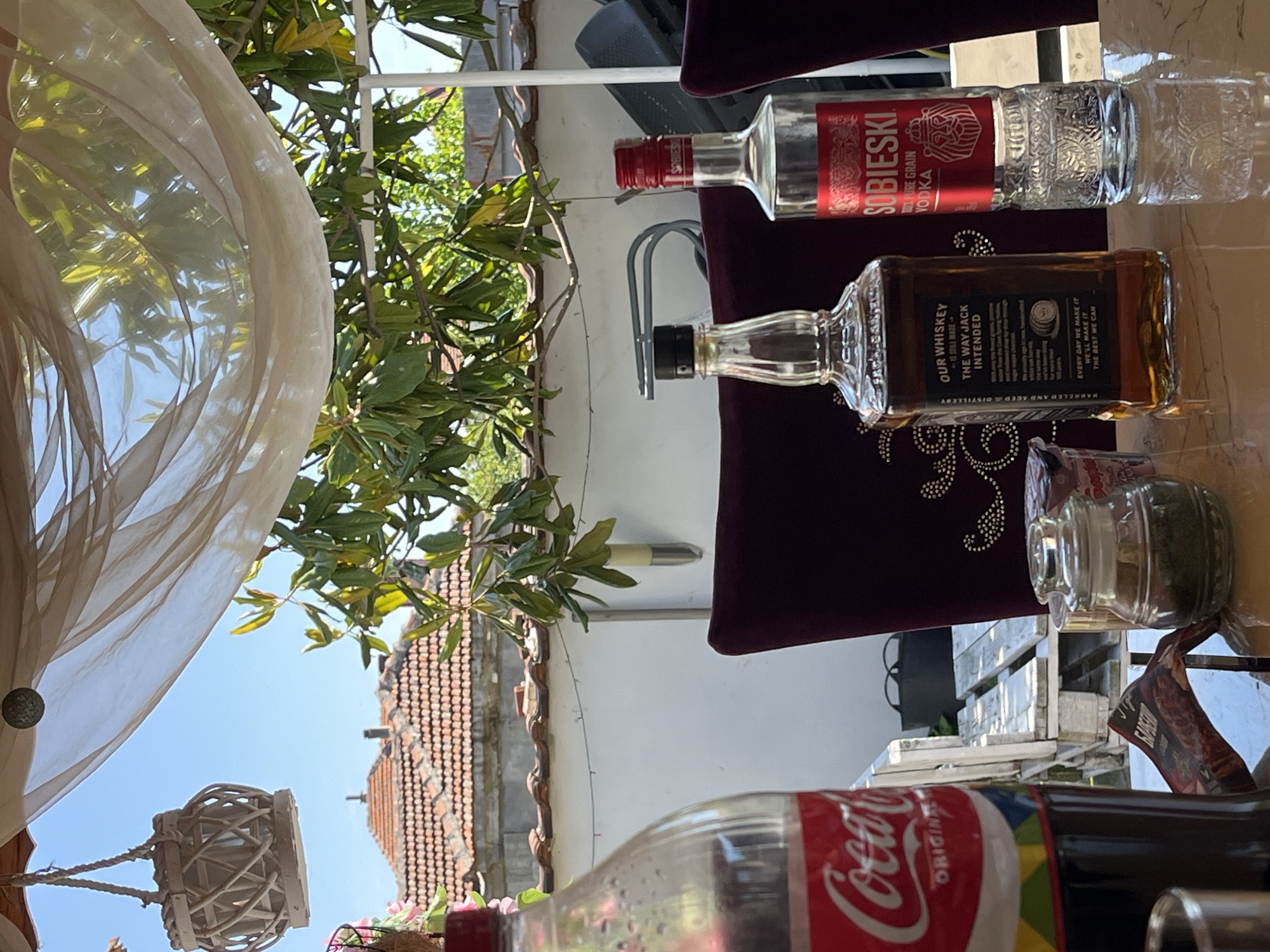 The image depicts an outdoor setting, possibly a patio or balcony, with a variety of objects placed on a table. A Coca-Cola bottle is visible in the foreground, along with bottles of Sobieski Vodka and Jack Daniel's Whiskey. A jar containing a green substance, possibly herbs or spices, is also present. A partially visible menu and seating hint at a restaurant or dining environment.
The background shows a tiled roof and foliage, suggesting a residential or semi-urban area. A beige sheer curtain hangs from above, adding a decorative element to the scene. The specific city and country are not discernible from the image.