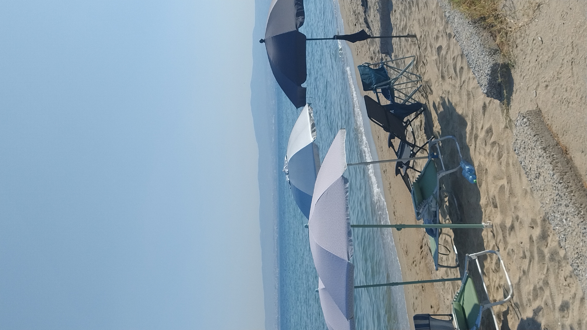 A sunny day at a beach in Perea, Greece. The scene features a sandy beach with the calm, blue waters of the Aegean Sea gently lapping the shore. Several beach umbrellas are set up, providing shade for empty lounge chairs. One large dark blue umbrella stands prominently on the right, with two lighter colored umbrellas to its left. Beach chairs, some folded and some in use, are scattered on the sand. A green and silver reclining beach chair is partially visible in the foreground. A blue plastic water bottle lies on the sand near the green chair. In the background, a hazy coastline with hills can be seen. The sky is clear and bright blue.