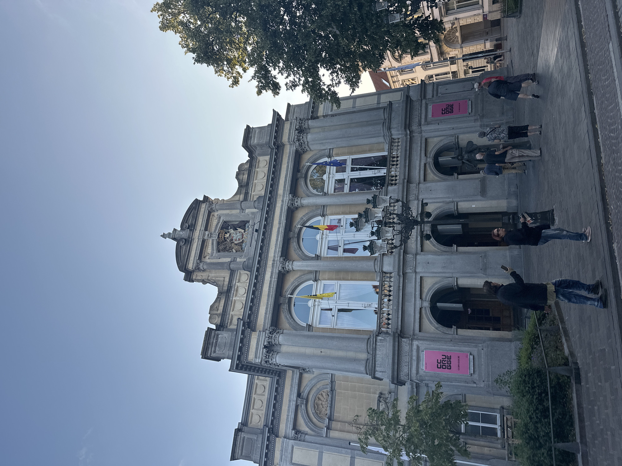The image shows a side of a grand, ornate building with neoclassical architectural elements. The building is made of light-colored stone and features columns, intricate carvings, and arched windows. Two flags are visible hanging from windows: one is the European Union flag, and the other appears to be a national or regional flag with yellow and red colors.
Several people are walking on the paved sidewalk in front of the building. They are casually dressed, suggesting a daytime scene. Some are looking at their phones, while others are engaged in conversation. The lighting indicates it is daytime, likely late afternoon due to the shadows. A large tree with green foliage is visible in the upper left corner of the image.
There are two prominent pink rectangular signs with white text on the facade of the building. One reads "CEU BRUGGE" and "CEUBRUGGE.BE". The text is upside down due to the orientation of the photo. The overall impression is a public space in a city, possibly a cultural institution or government building, in Bruges, Belgium. The atmosphere appears relaxed and ordinary.