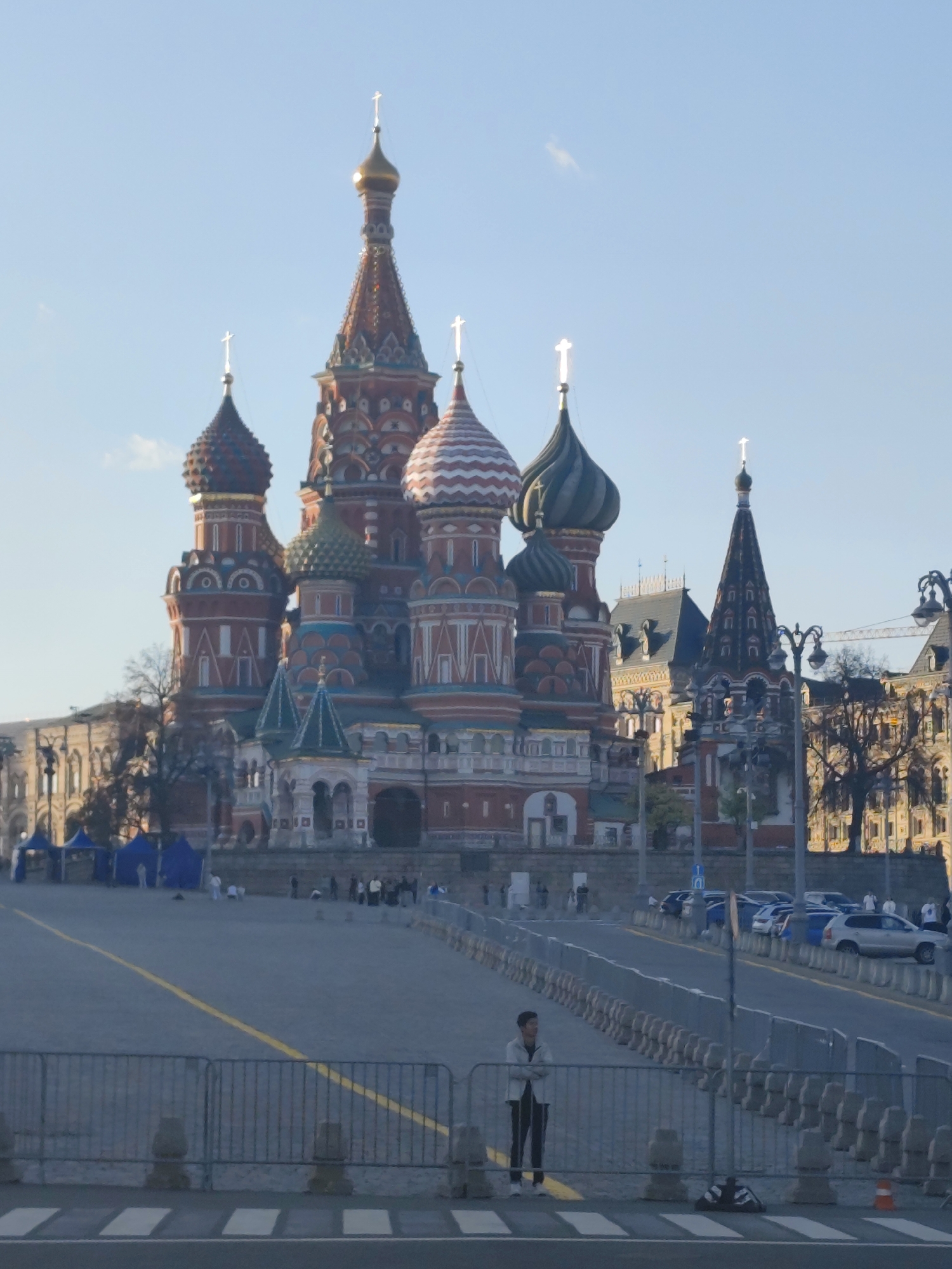 The image depicts St. Basil's Cathedral in Moscow, Russia, under a clear, blue sky. In the foreground, a metal fence runs across the frame with a pedestrian visible on the street behind the fence. Behind the fence, a vast paved area stretches toward the cathedral. Some people stand or walk in the distance. In the background to the right of the cathedral, other buildings are visible. Several cars are parked on the street that runs along the edge of the paved area. Blue tents are visible in the distance near the left of the paved area.