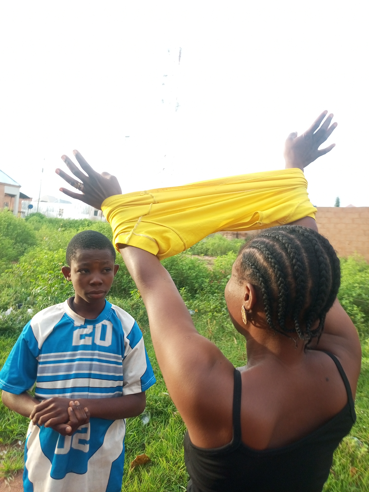 In Tempure, Nigeria, a young boy stands on a grassy area with a building and lush vegetation in the background. He is wearing a blue and white shirt with the number 20 visible on the front. To the right, an adult woman with braided hair is holding up a yellow garment with her hands. Her arms are raised above her head, and the yellow fabric is draped over them. The sky is bright and overcast.