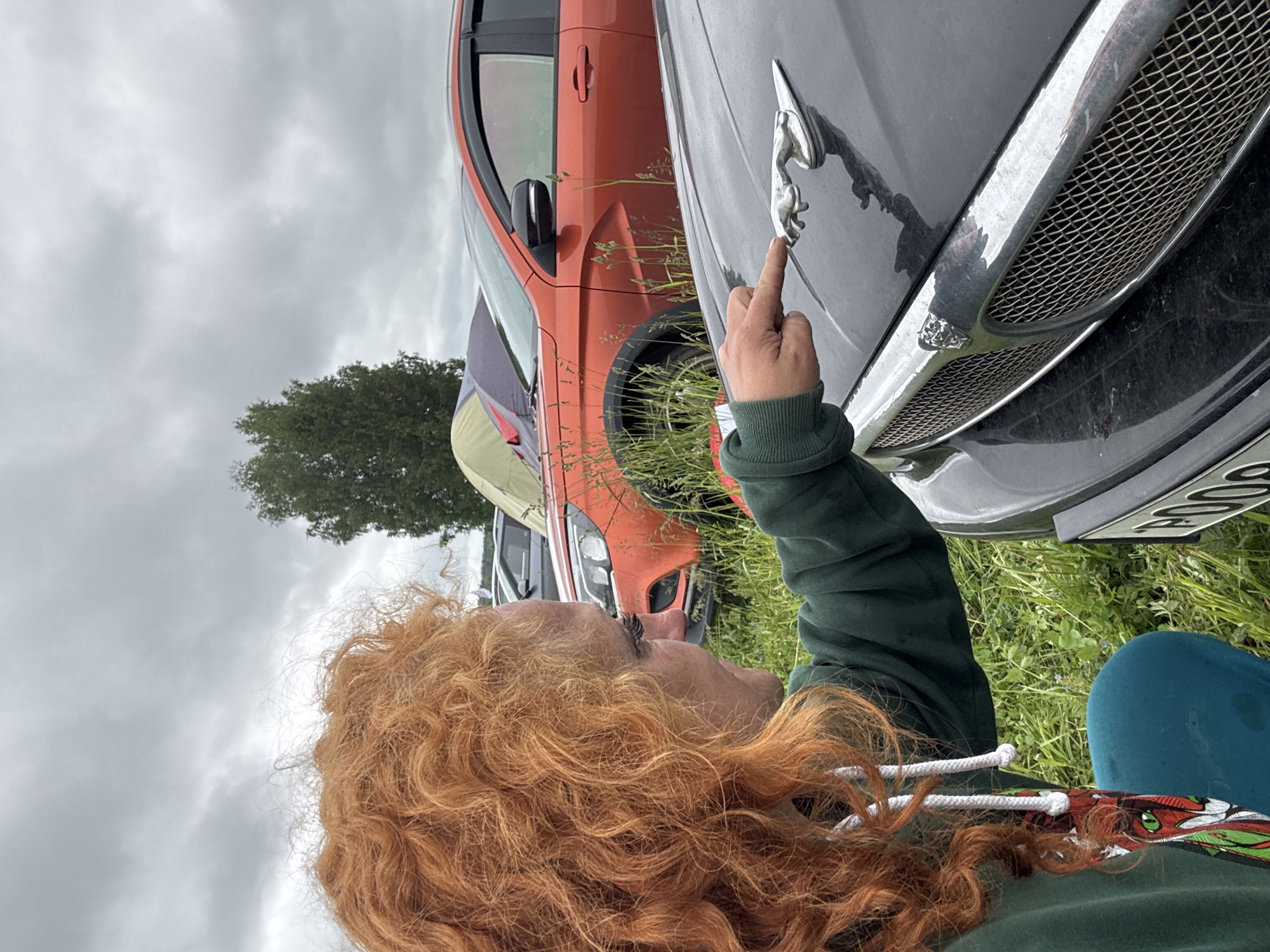 The image depicts an outdoor scene in Большая Кивара, Russia, under an overcast sky. In the foreground, a person with voluminous, curly red hair, wearing a dark green hooded sweatshirt with white drawstrings and teal-colored trousers, is in a crouching position. Their right arm is extended, with the index finger pointing directly at a chrome "leaping jaguar" hood ornament on the front hood of a dark grey or black car.
The car features a prominent chrome mesh grille with a smaller Jaguar head emblem centered on it. A partially visible Russian license plate, beginning with "P00," is affixed to the lower front bumper. The car's hood surface shows minor dust or dirt.
In the mid-ground, a vibrant orange-red crossover SUV with black fender flares and tinted windows is parked in tall green grass. Behind it, a dark grey vehicle and at least two camping tents are visible, suggesting a temporary outdoor gathering or campsite. One tent is light green, and another darker tent is behind it. A tall, dark green coniferous tree stands on the right side of the background. The ground is covered in dense green grass and various weeds.