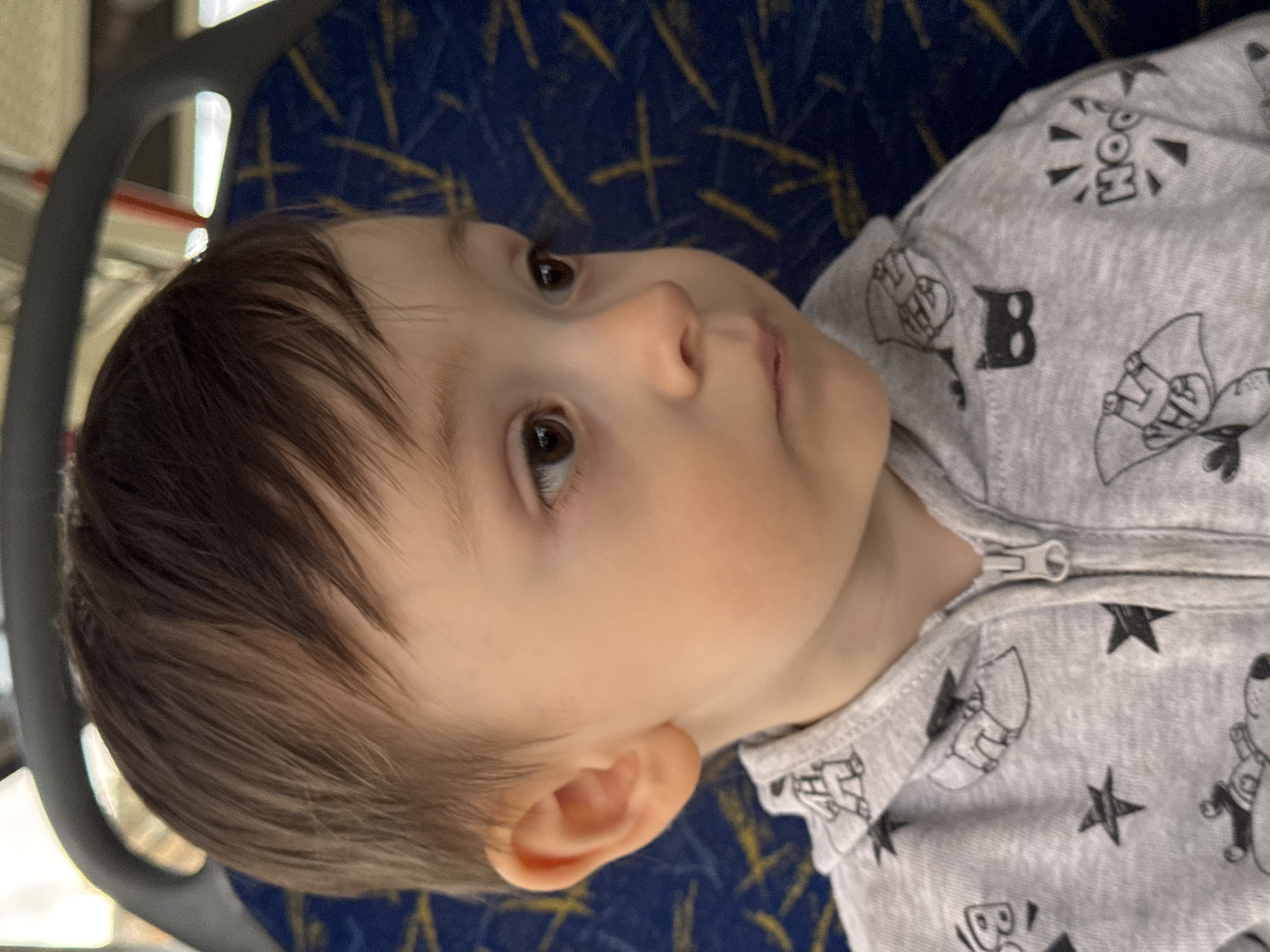 A close-up, head-and-shoulders portrait of a young child, likely a boy, with brown hair and brown eyes. The child is wearing a grey hooded sweatshirt with a zipper. The sweatshirt is adorned with cartoonish black and white graphics, including stars, comic book-style speech bubbles with the word "HOO!", and caped figures resembling superheroes. The child is looking to the right, with a neutral expression. The background is a blurry, dark blue and yellow patterned fabric, suggestive of seating upholstery. The overall lighting indicates an indoor environment, possibly public transportation, with natural light filtering in from the left. The image provides no specific geographical indicators to determine its location within the city of Izhevsk, Russia.