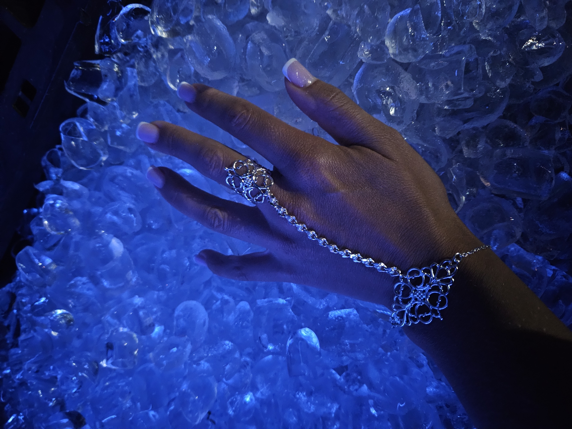 A close-up shows a dark-skinned hand adorned with a silver filigree hand chain and ring, resting on a bed of ice. The hand has well-manicured nails with a French manicure. The ice is illuminated with a blue light, giving the scene a cool, almost surreal atmosphere. The location is Unknown, Unknown.