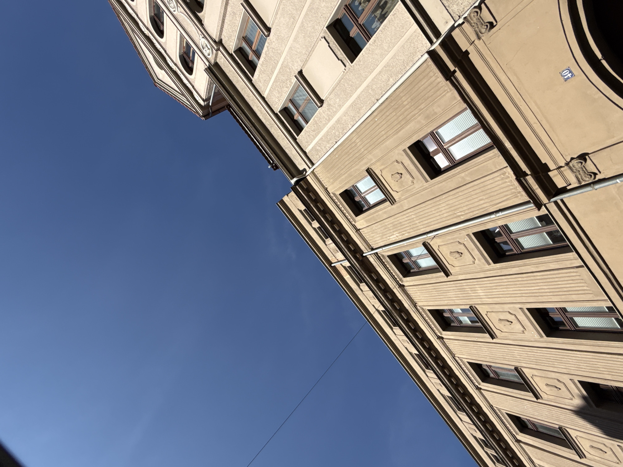 The image captures a low-angle view of two tan-colored buildings against a clear blue sky, suggesting a daytime scene in Görlitz, Germany. The building in the foreground, with ornate detailing and multiple windows, is angled to the left. Its facade features vertical ribbed sections and decorative rectangular insets, each containing a stylized emblem. The windows are framed with dark brown wood and some are fitted with white blinds. The building on the right, seen at a higher elevation and angled to the right, has a similar architectural style with arched windows visible on its upper floors. A single black wire stretches diagonally across the upper portion of the frame, cutting through the sky. The number "40" in a blue square is visible on the lower section of the foreground building, likely indicating a street address. The overall impression is one of historic European architecture under a bright sky.