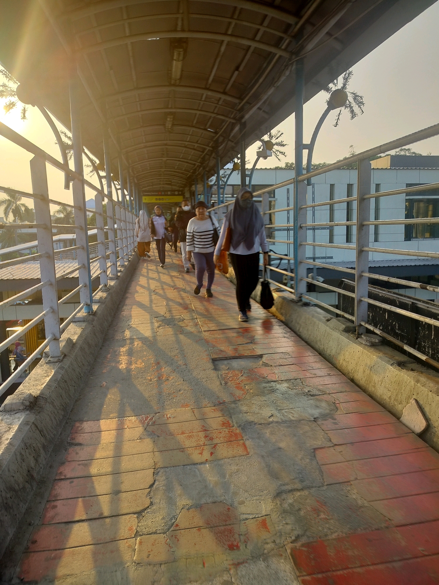 This image depicts a covered pedestrian walkway, likely an overpass, on a sunny day in East Jakarta, Indonesia. The walkway is constructed with a metal roof supported by a blue metal frame and features a railing on either side. The floor of the walkway is a mix of concrete and red-colored material, possibly brick or painted wood, with some areas showing wear and tear.
Several people are walking along the walkway. In the foreground, a woman wearing a dark hijab and black pants is walking away from the camera, carrying a black bag and an orange bag over her shoulder. Further ahead, a group of people are walking in the same direction. One woman wears a striped sweater and dark pants, another is wearing a light-colored hijab and pants, and a person in the middle is wearing a face mask. To the left, a woman in a white top and patterned skirt is visible. The sunlight creates long shadows across the walkway, indicating it is either early morning or late afternoon.
The background shows a building with a modern facade, characterized by horizontal window elements, and some palm trees, suggesting a tropical climate. The overall environment appears to be a public space, possibly connecting two areas of a building or providing access over a road or railway. The presence of a sign with yellow text in Indonesian is visible on the ceiling of the walkway, indicating this is an official public thoroughfare.