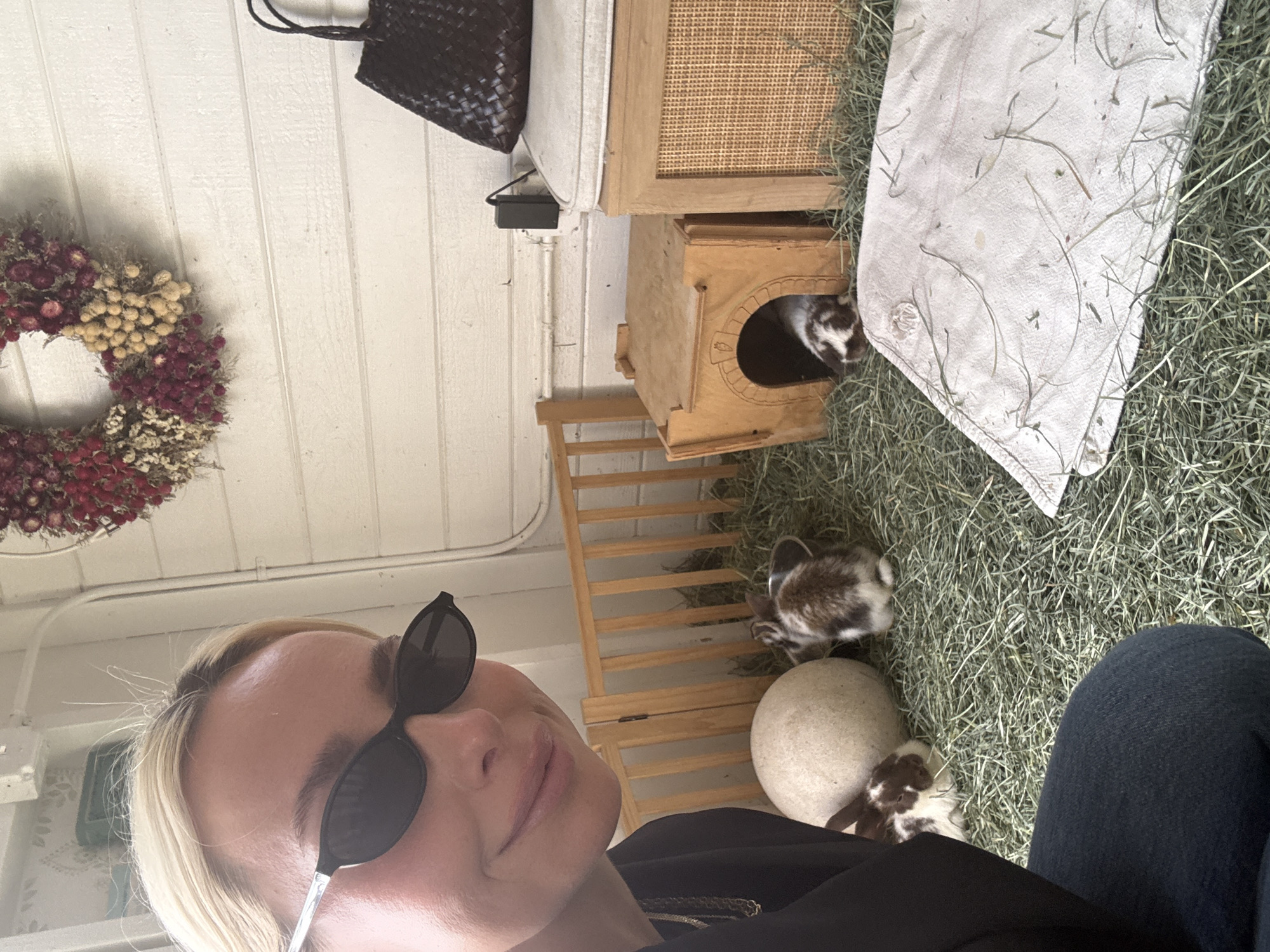 The image shows a person with blonde hair and sunglasses taking a selfie indoors with rabbits. The background includes a white-painted wall with a floral wreath, a woven black bag, a wooden cabinet, a small wooden house with a rabbit peeking out, and what appears to be a rabbit enclosure filled with hay. The environment suggests a pet-friendly or animal-related setting. There is a stone-like orb near a rabbit and a partial view of a person wearing jeans. I have no evidence of the location in the city of Solana Beach, United States of America.