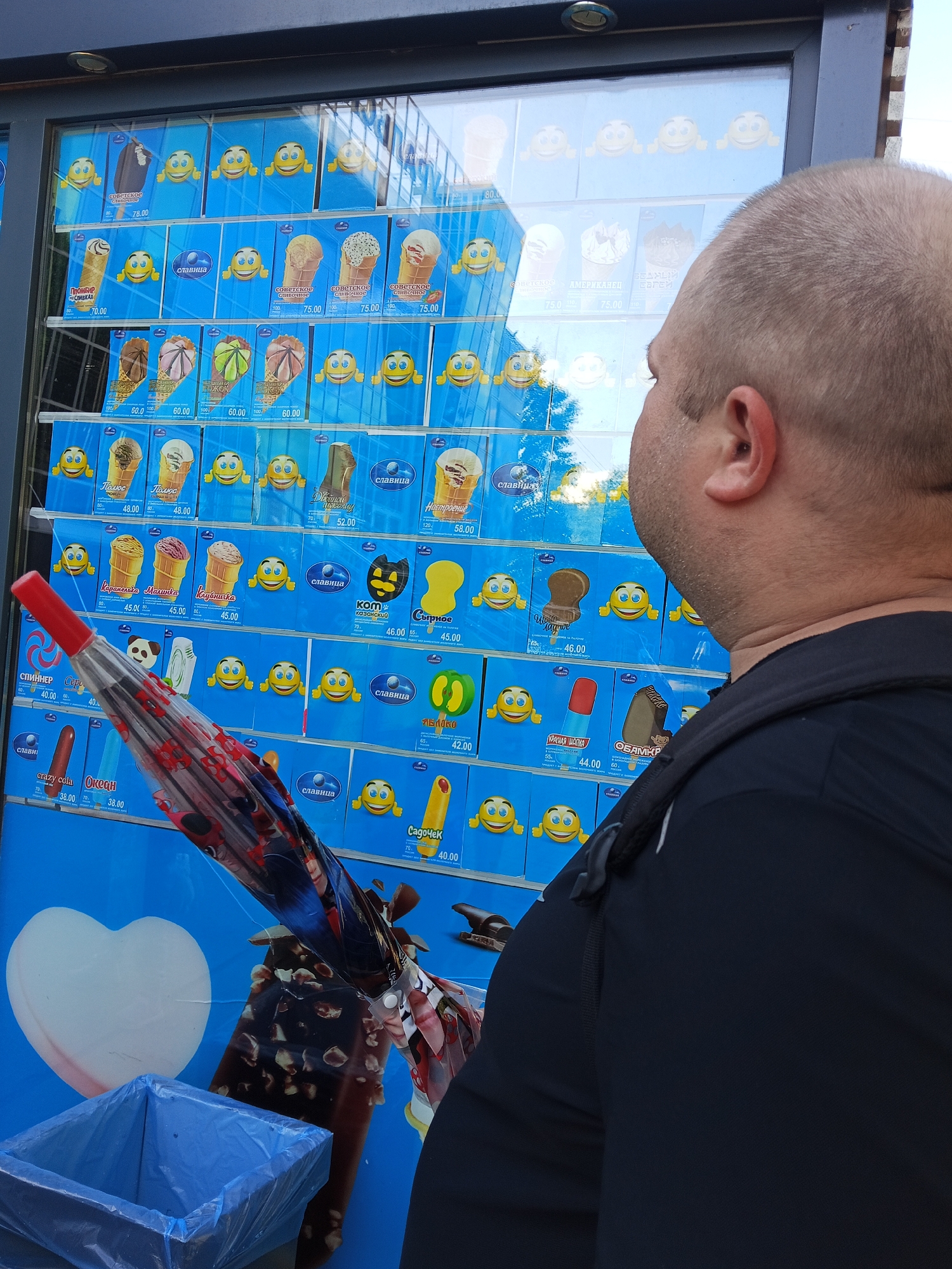 In Chaikovsky, Russia, a man wearing a black t-shirt and backpack is looking at an outdoor ice cream display. The display features numerous ice cream options, each with a picture of the product, its name, and price. The background shows a reflection of trees and buildings. A partially opened umbrella with a cartoon design is held in front of the display. To the left of the ice cream display, a blue trash bin is visible. The man appears to be considering his purchase.