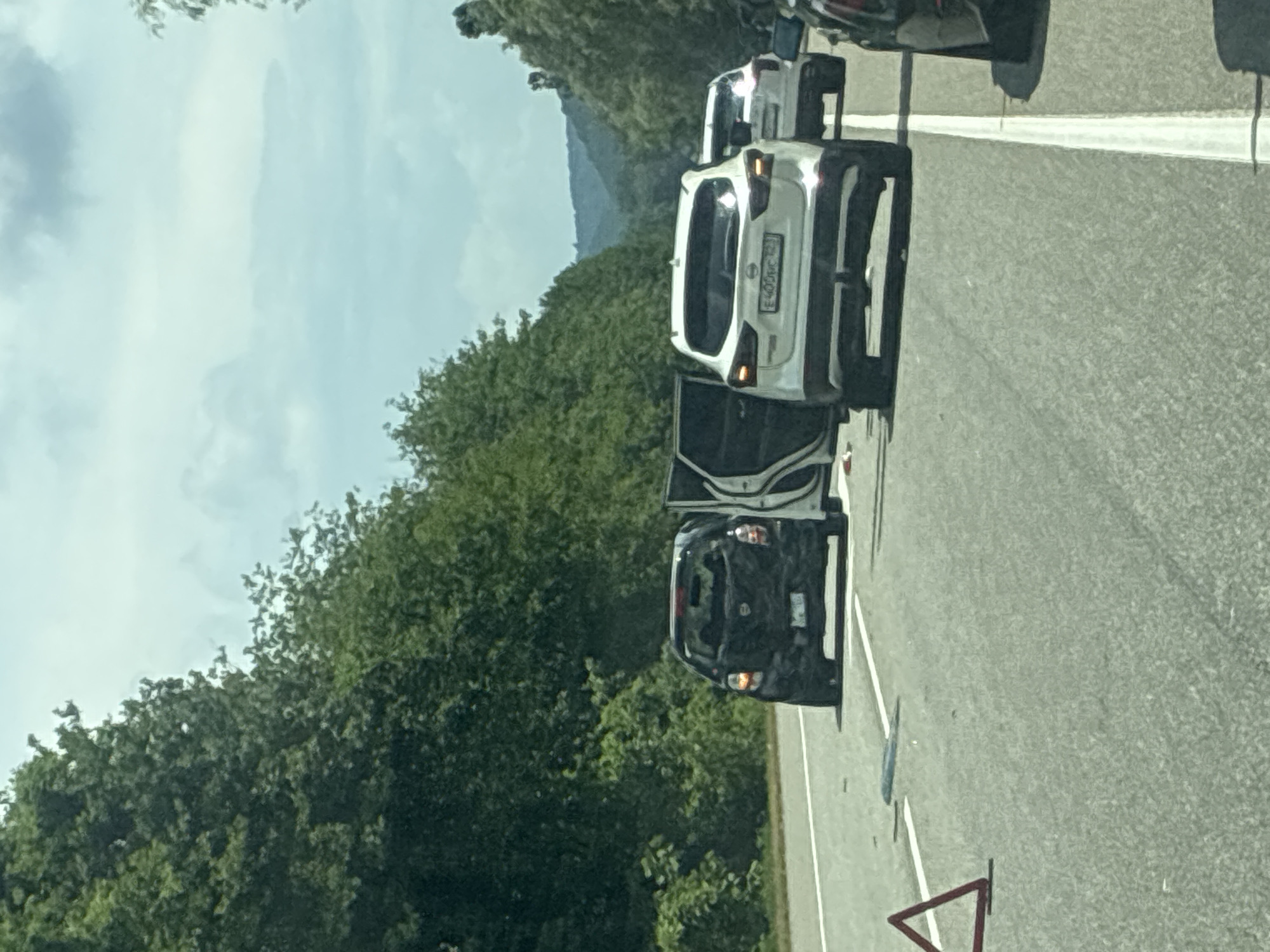 The image depicts a traffic incident on a highway, possibly in or near Tekos, Russia, characterized by a line of cars stopped on the asphalt. The foreground shows the right side of a highway with white lane markings. A red triangular warning sign is visible on the left side of the road, indicating a hazard.
Several vehicles are present. A black hatchback with its rear facing the camera is on the left side of the road, its driver's side door and rear passenger door are open. To its right, a white SUV is positioned, its rear facing the camera, with its driver's side door open. Behind the white SUV, another white car is visible, also stopped. On the far right, the back of a dark-colored car is partially visible.
The surrounding environment is a highway bordered by dense green trees on both sides, with hills or mountains in the background under a cloudy sky. The lighting suggests it is daytime. The presence of open car doors and the warning sign suggests a possible accident or breakdown has occurred. No people are clearly visible in the image.
