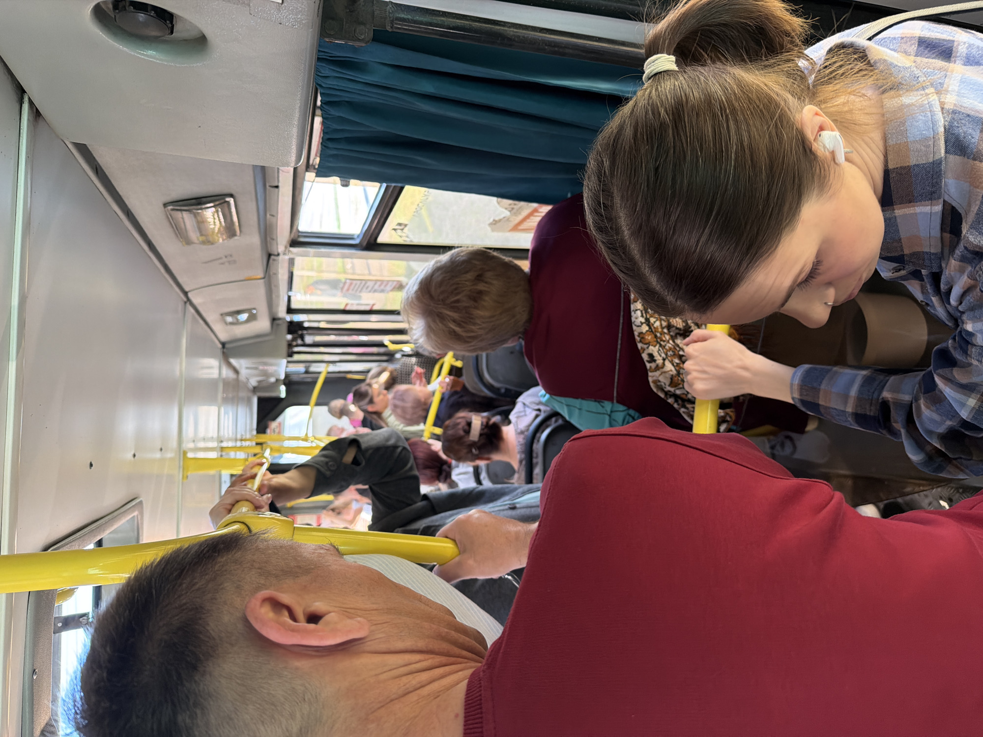 The image shows the interior of a crowded bus, likely during transport. A diverse group of people is present, holding onto yellow handrails for support. One woman with her hair in a bun and wearing a plaid shirt is looking down, her attention seemingly focused on something near her. An older man with short hair and a red shirt is also holding onto a handrail, his face in profile. The bus has a blue curtain. Given the prompt to describe the location, this likely occurred on a bus in Krasnoyarsk, Russia.