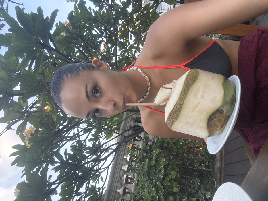 A female subject is depicted outdoors in Bangkok, Thailand, looking directly into the camera while drinking. Her dark hair is pulled back, revealing a fair complexion. She wears a dark gray bikini top with neon orange trim and a white cowrie shell choker necklace. Her lips are pursed around a light-colored straw inserted into a partially husked young coconut, which rests on a small white plate. A maroon fabric is draped over her lower body. The background features lush green Plumeria trees with light yellow blossoms, decorative string lights, a white classical-style balustrade, and other green foliage, indicative of an elevated outdoor terrace or garden environment with wooden decking.