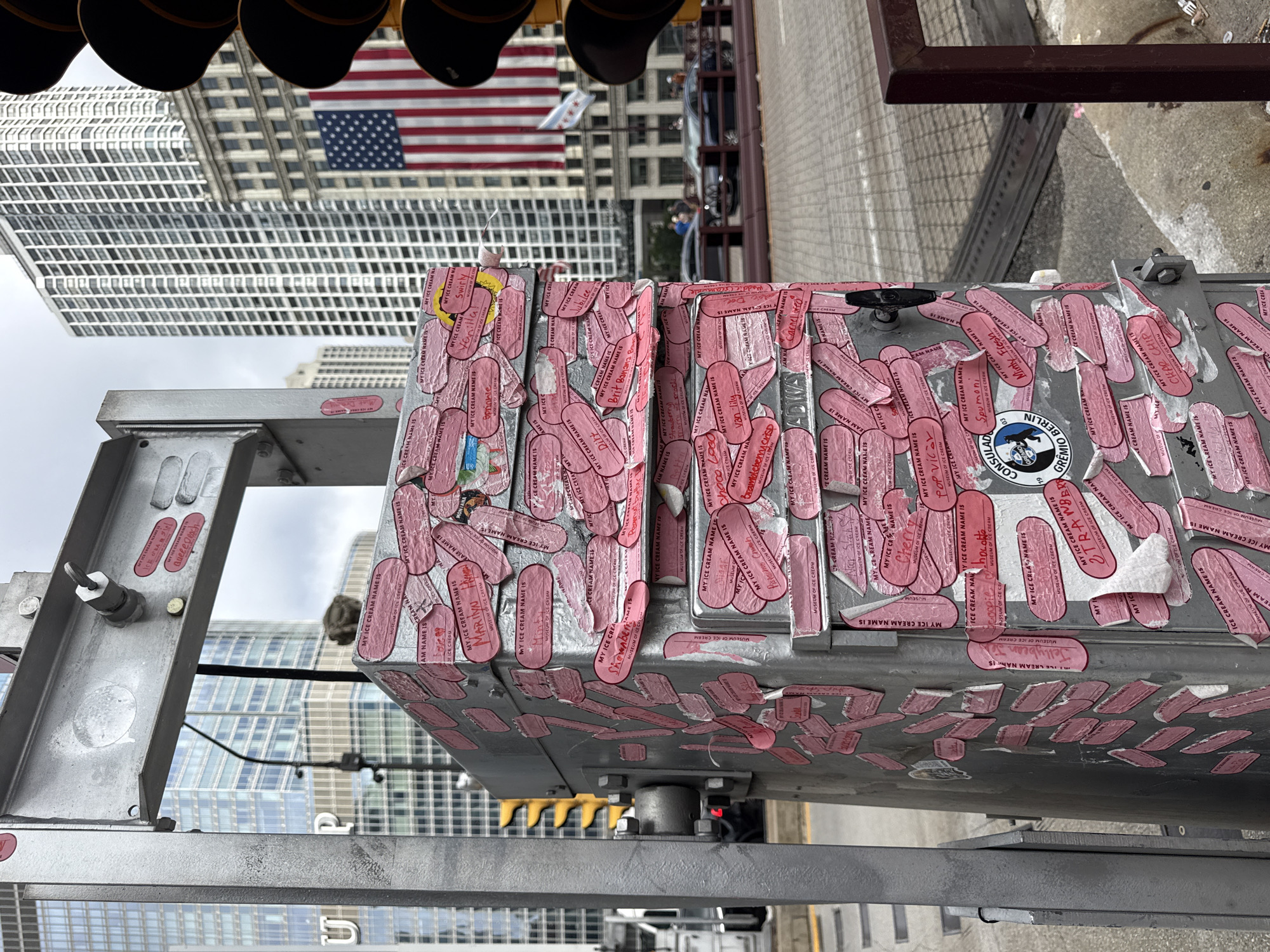 This media file depicts a close-up view of a metal utility box covered in numerous pink stickers, each bearing the text "MY ICE CREAM NAME IS" followed by a unique name and "MUSEUM OF ICE CREAM." The box is positioned outdoors, with a cityscape visible in the background. In the upper portion of the image, the American flag is prominently displayed, suggesting a location in the United States, further confirmed by the "CHICAGO" and "GREMIO BERLIN" text on some stickers.
The overall scene suggests a public art installation or a collaborative project, possibly related to the "Museum of Ice Cream." The stickers appear to be a collection of ice cream flavor names or personal ice cream-themed monikers submitted by visitors. The metal box itself appears to be part of some outdoor infrastructure, possibly traffic control equipment given the presence of what looks like traffic light lenses at the very top. The background shows modern skyscrapers, indicating an urban environment. The lighting suggests it is daytime, possibly overcast or late afternoon, as there are no harsh shadows. The image is taken from a slightly elevated, slightly tilted perspective. The sheer volume of stickers implies the project has been ongoing for some time or has been very popular.