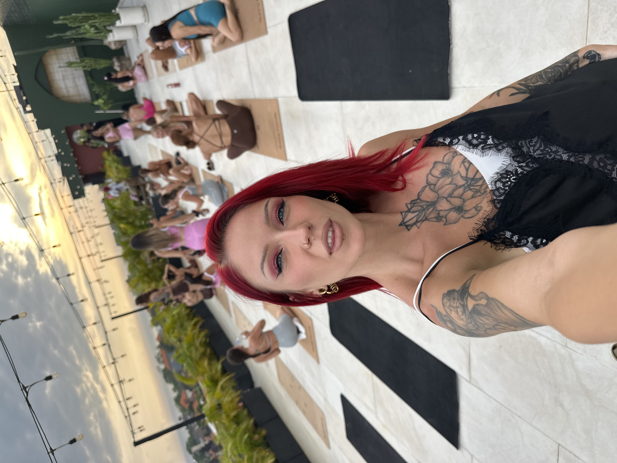 The image is a selfie taken outdoors, likely in Tibubeneng, Indonesia, based on the visible skyline and greenery. The woman in the foreground has red hair, visible tattoos on her chest and arm, and is wearing a black top with lace detailing. Behind her, a group of individuals are positioned on yoga mats, presumably participating in a yoga session. The yoga mats are placed on a striped tiled surface. In the background, there's greenery, a building with a visible window, and the skyline, suggesting an elevated location. The lighting indicates it's either sunrise or sunset. Street lamps are also visible in the background.
