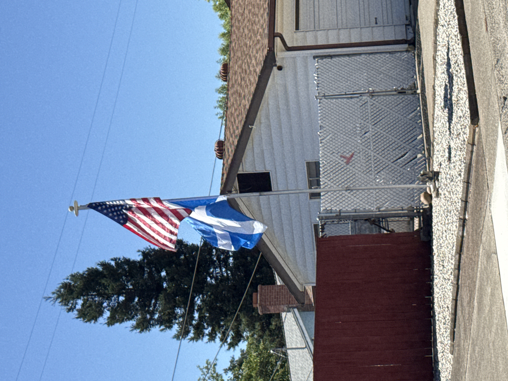 The image shows two flags flying on a flagpole in front of a house. The top flag is the flag of the United States of America, and the bottom flag is the flag of Scotland. The house is a single-story, light-colored structure with a brown shingled roof and a brick chimney. There is a red wooden fence in the foreground to the left of the flagpole, and a chain-link fence behind it. A large evergreen tree is visible to the left of the house, and power lines are strung across the sky. The scene appears to be a residential area. The location is identified as Citrus Heights, United States of America.