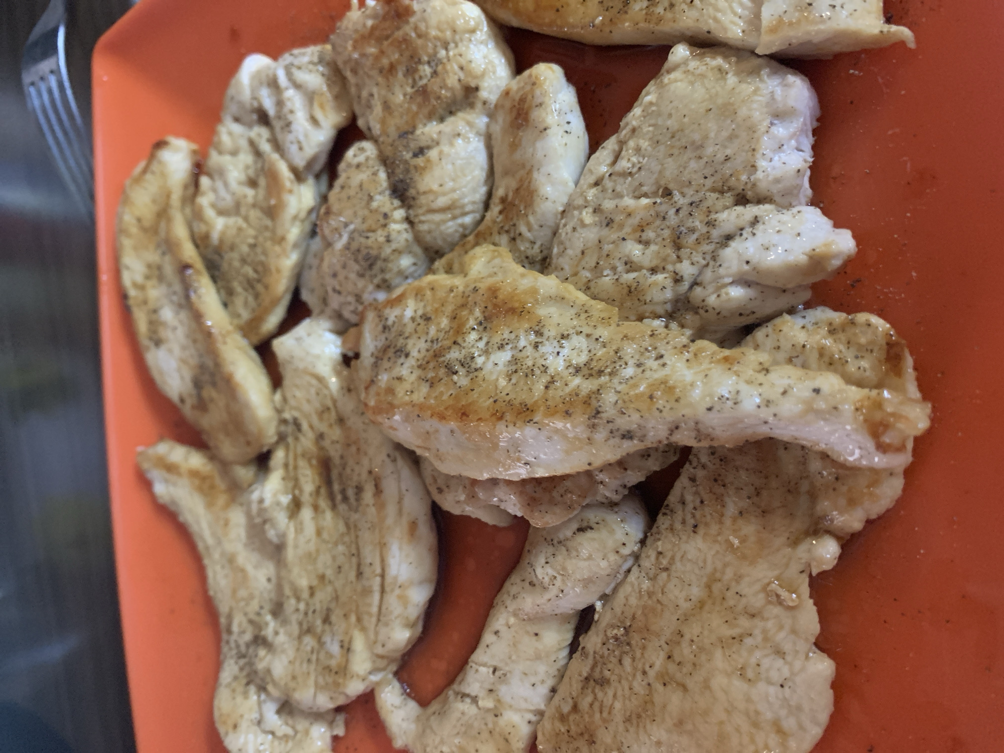 The image displays a close-up, overhead view of cooked chicken pieces served on an orange, rectangular plate. The chicken appears to be cut into irregular strips or tenderloins, ranging in color from off-white to light golden brown, indicating they have been cooked, possibly pan-fried or baked. Many of the chicken pieces are seasoned with visible specks of black pepper. Some pieces show slight browning and a glossy sheen from cooking juices or oil.
The orange plate occupies most of the frame, providing a vibrant backdrop for the food. In the top-left corner, the tines of a silver metal fork are partially visible, suggesting the chicken is ready to be eaten or has just been served. The lighting suggests an indoor setting, possibly a kitchen or dining area. No people are visible in the frame. The overall scene depicts a simple, prepared meal. The geographical context provided for this image is Vozdovac Urban Municipality, Serbia. There is no visible text in the image.