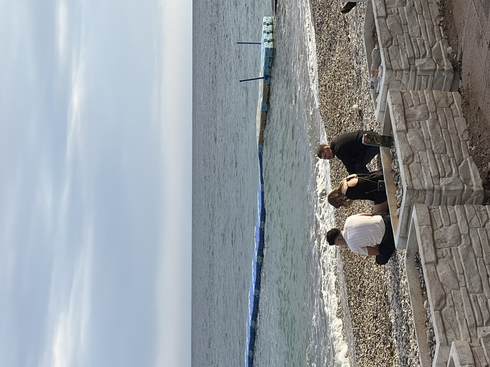The image depicts a scene at a pebble beach in Budva, Montenegro, on an overcast day. Three individuals, two women and one man, are at the edge of the beach. A man is wearing a white shirt and black pants, a woman has black dress, and the third person is a man wearing a black top. They appear to be looking at the beach pebbles and searching for something. A long, blue breakwater stretches parallel to the shoreline, situated partly in the water and partly on the pebbles. A concrete wall with stone-like sections stands adjacent to the beach. Litter is seen on the wall. The sky is grey.