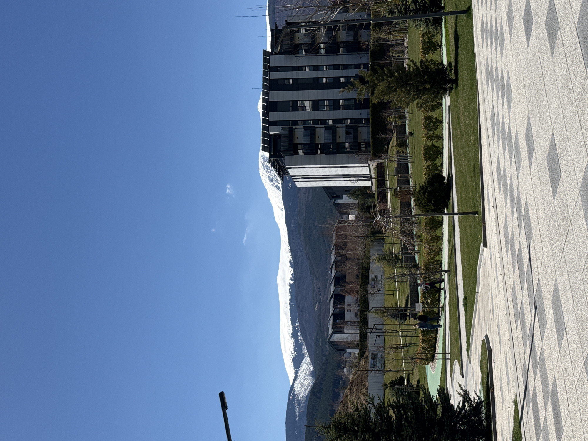 The image shows a view in Sofia, Bulgaria. In the distance, a snow-capped mountain is visible. Closer to the camera is a modern, tall building with a black and white facade. In front of the building is a park with green grass, trees, and walkways. A paved area with a diamond pattern design is in the foreground. A lamppost is visible in the lower-left corner. No people are clearly identifiable. The sky is bright blue.