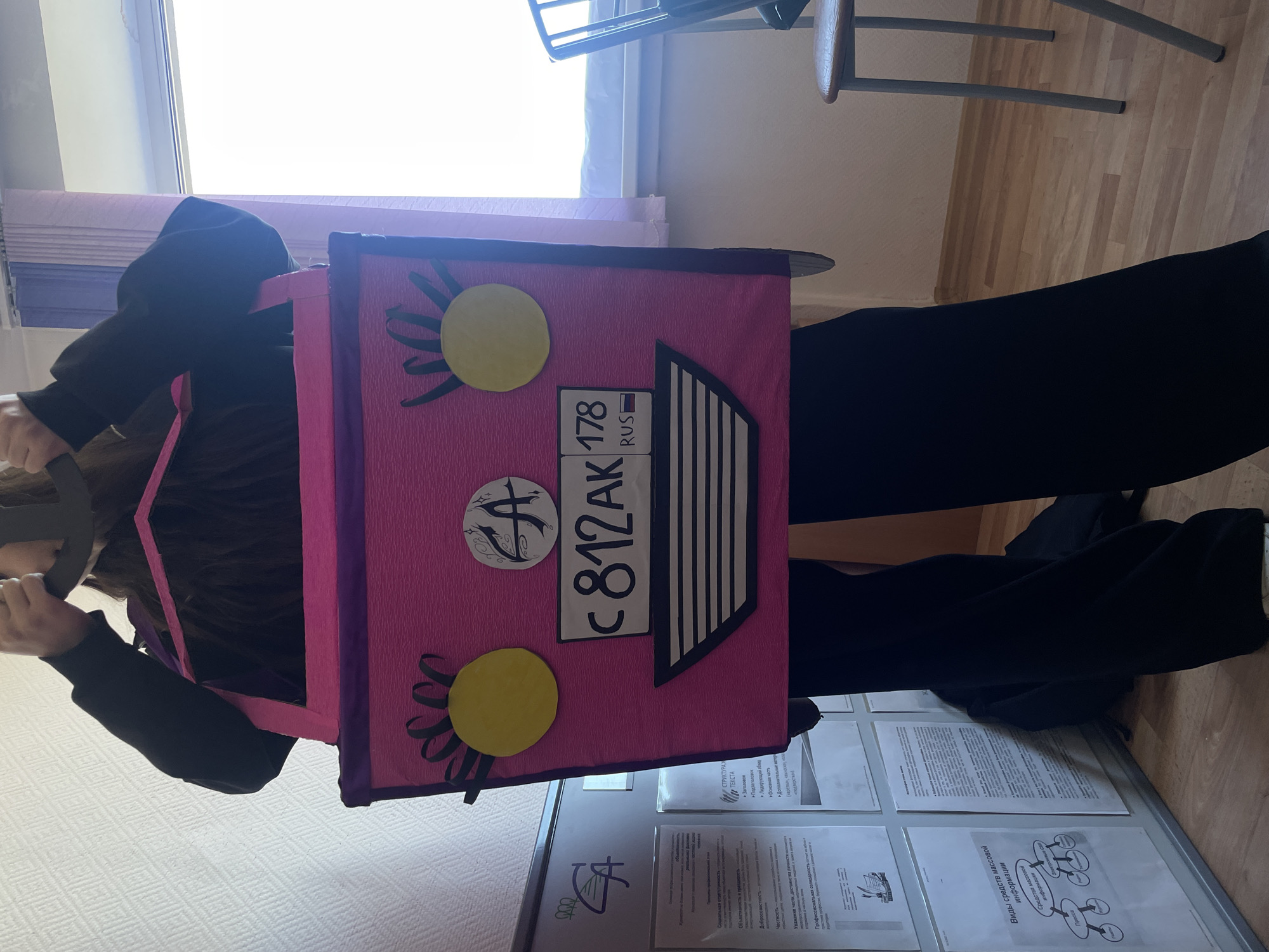 A person wearing black clothing is carrying a rectangular, pink prop with a stylized license plate that reads "C 812 AK 178 RUS". The prop also features yellow circles with black curly decorations. The person's face is partially obscured. The background includes a window, a chair, a wall with posters, and a wooden floor. Based on the "178 RUS" code on the license plate, this scene is likely located in Saint Petersburg, Russia.