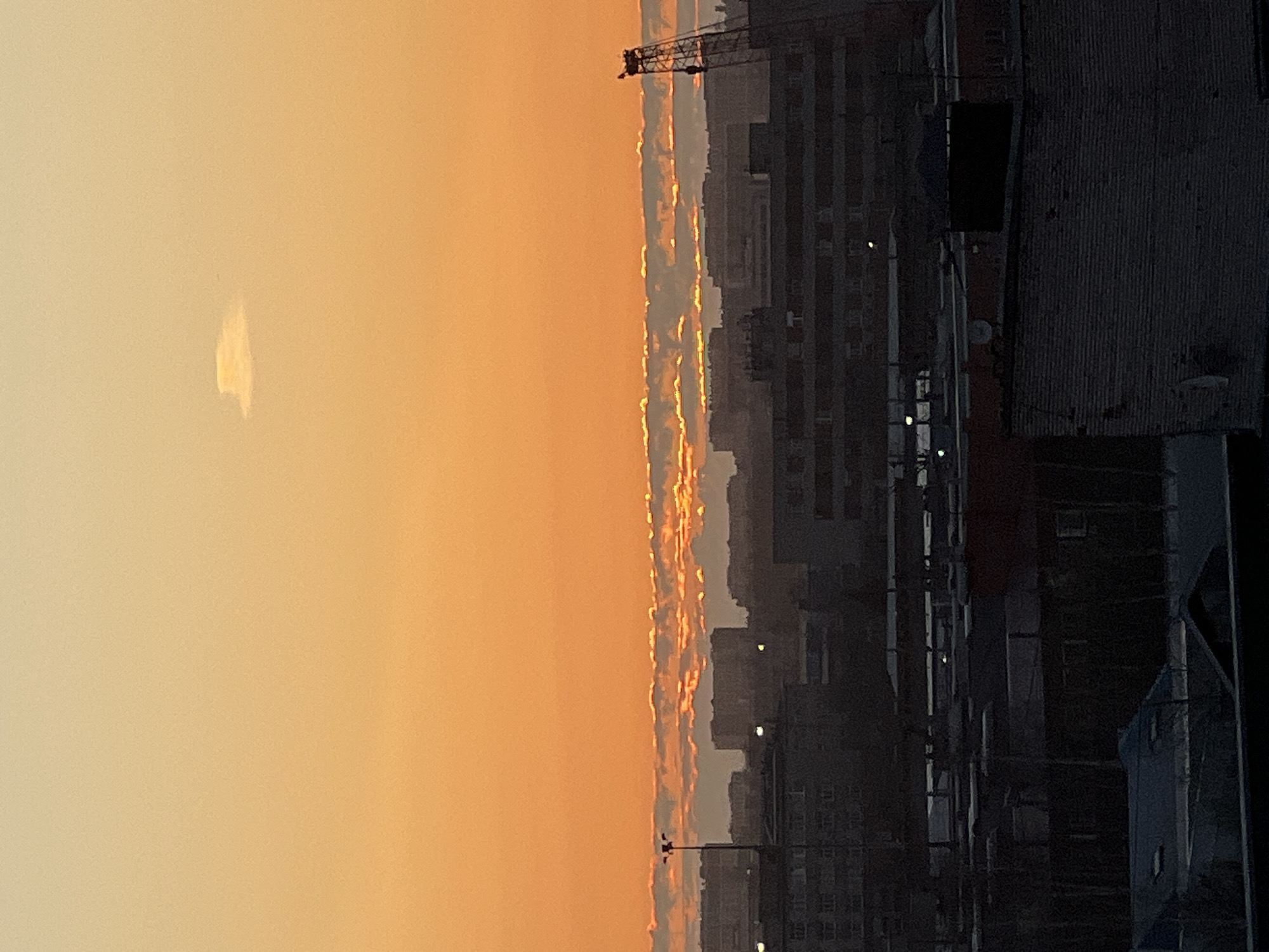 The image depicts a sunset over a cityscape in Saint Petersburg, Russia. The sky transitions from a pale yellow to a vibrant orange. A cloud is visible on the left side of the image. The right side of the image is filled with buildings, with visible windows and rooftops. A crane is visible in the skyline. The image is rotated 90 degrees counter-clockwise.