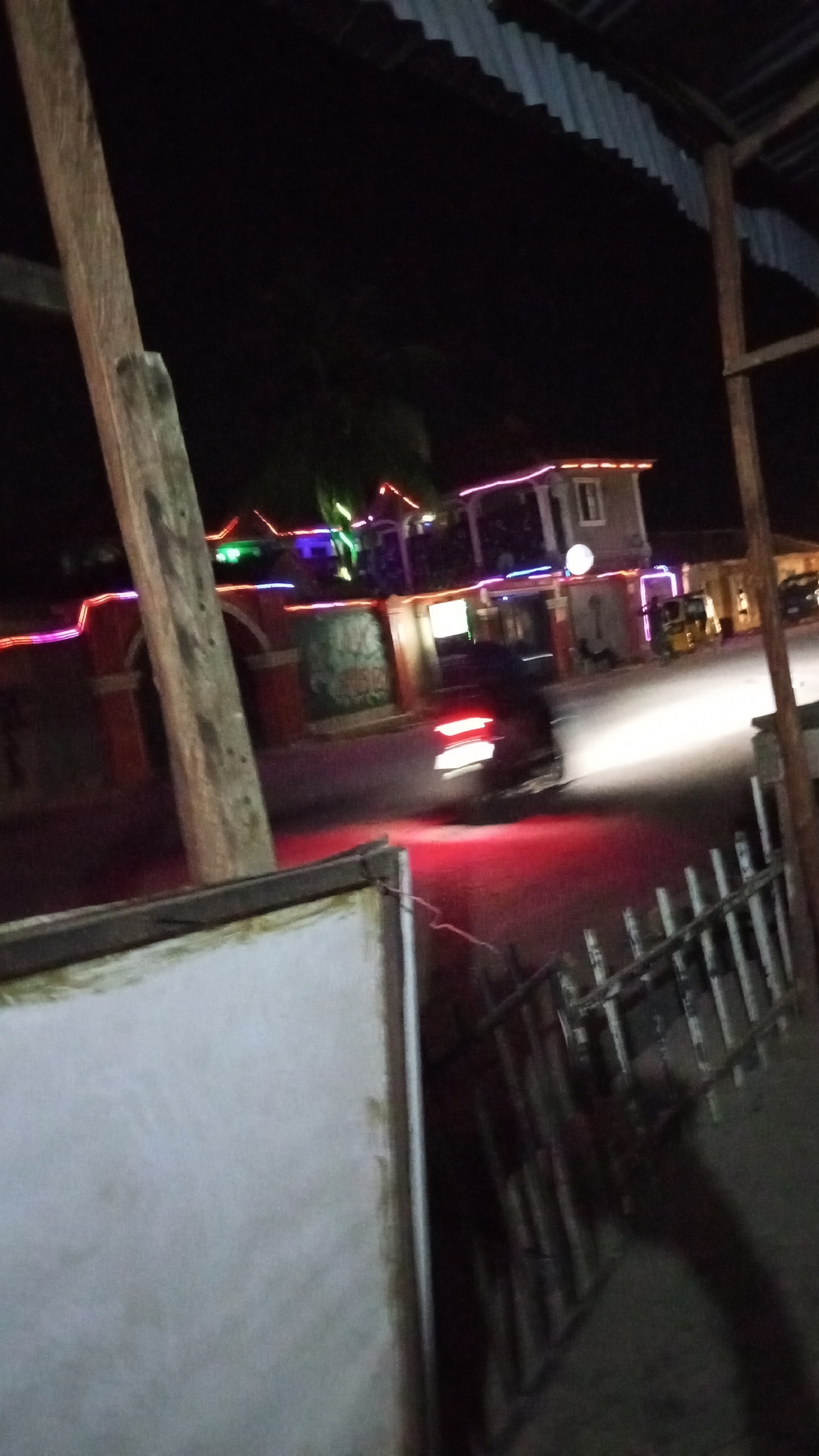 The image, taken at night in Sabon Tasha, Nigeria, shows a street scene with blurred moving headlights and taillights of a vehicle. Buildings decorated with colorful neon lights line the street. A tricycle is visible parked on the side. In the foreground, a weathered wooden post and a section of a white structure and a fence obstruct the view. A corrugated roof overhangs the scene at the top of the image.
