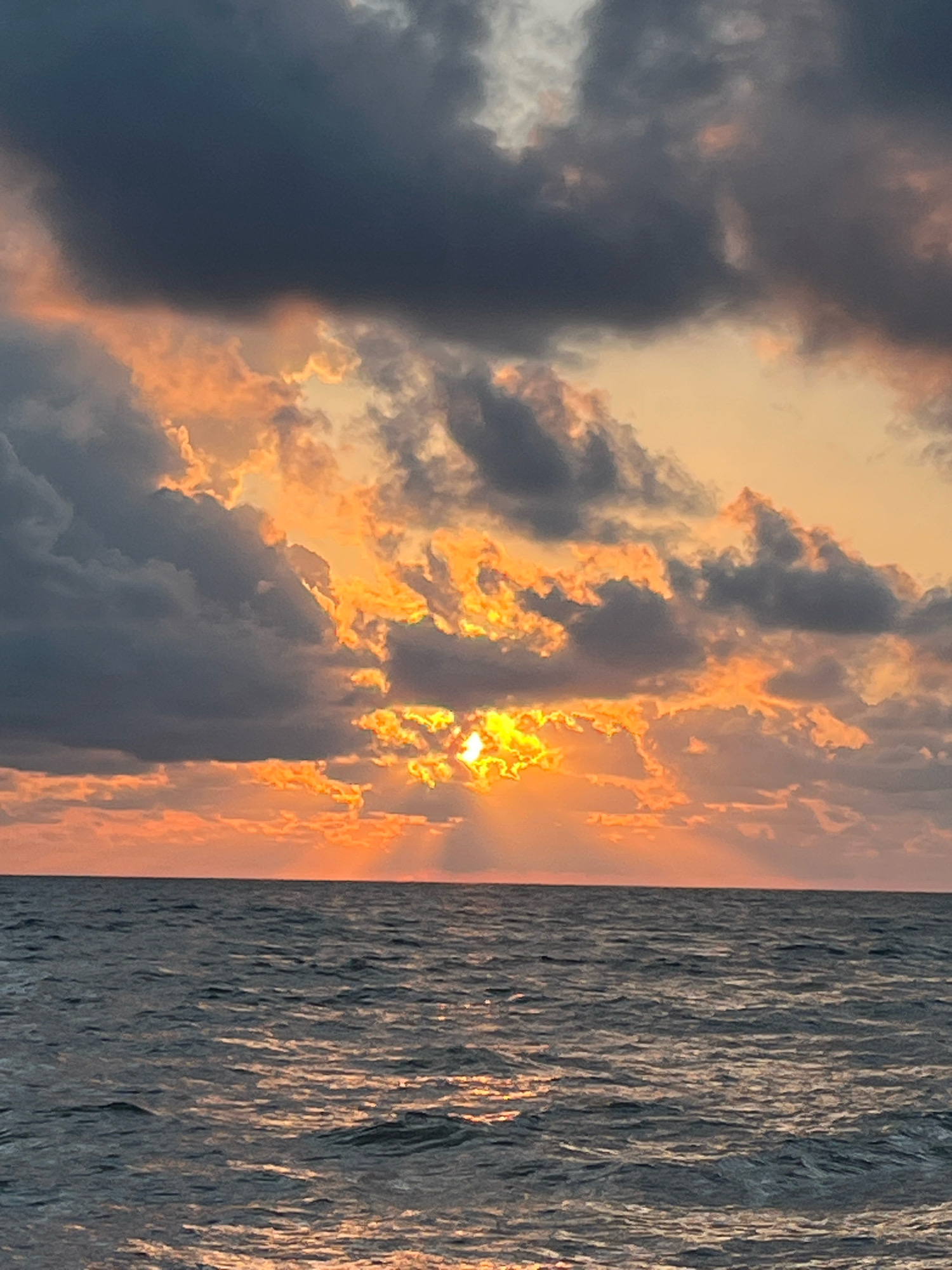 A sunset over the Black Sea in Ureki, Georgia, features dark, textured clouds illuminated by the setting sun. The sun's rays pierce through the clouds, casting an orange glow and creating a dramatic contrast with the darker portions of the sky. The sea below is choppy, reflecting the colors of the sunset, with the horizon line clearly visible.