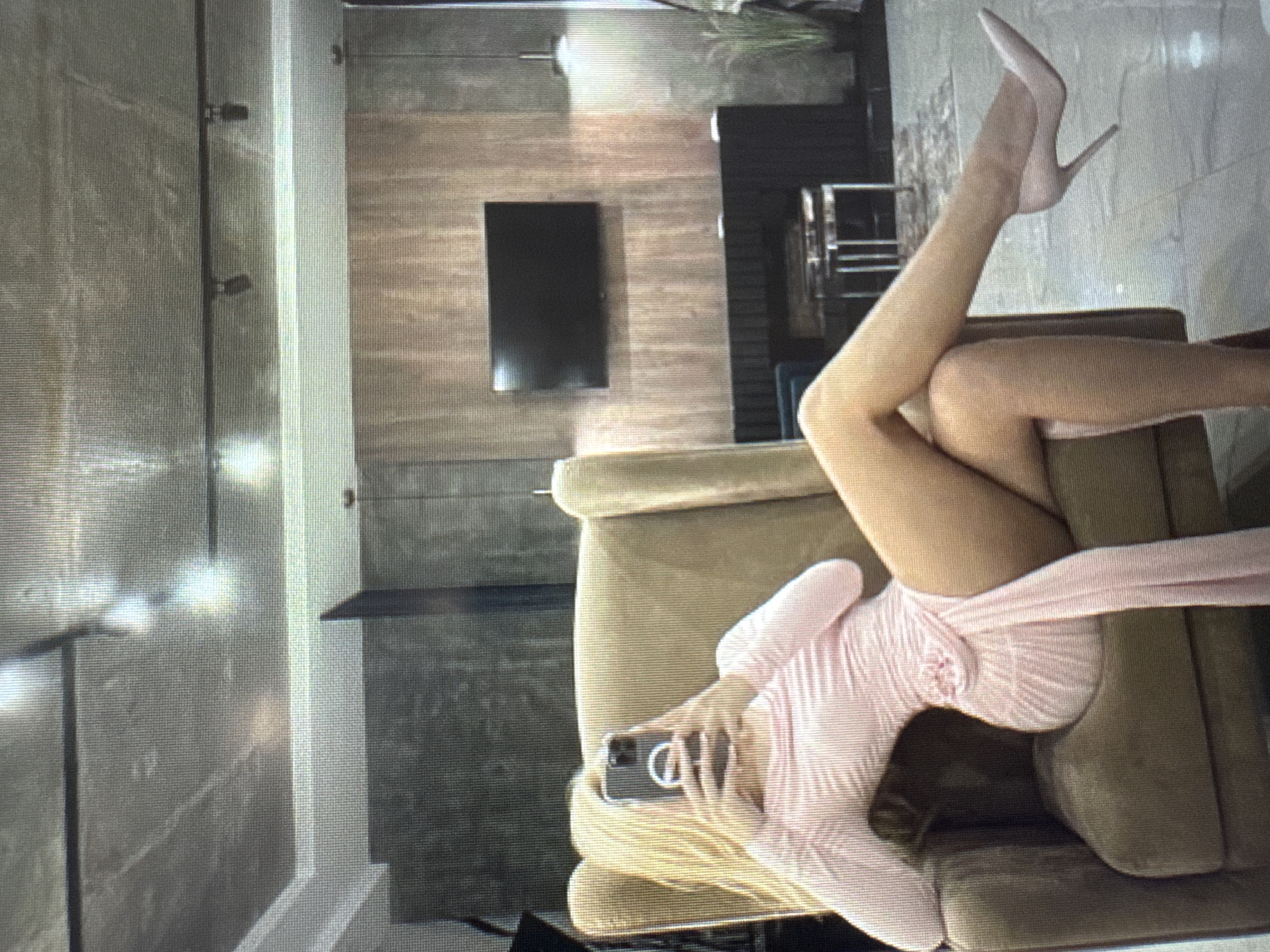 A person is lying horizontally across a light brown upholstered chair or sofa, positioned centrally in the image. They are wearing a light pink long-sleeved garment, possibly a dress. One leg is extended upwards, wearing a light pink high-heeled pump, while the other leg is bent. The person is holding a smartphone in both hands near their chest. The smartphone has a clear case with a prominent white, circular or 'D' shaped design on its back.
The background depicts a modern indoor room. On the left side of the frame, there is a wall with horizontal wood paneling, on which a dark, rectangular wall-mounted television screen is visible. To the right of the wood paneling, another lighter-colored wall or pillar is present. Further into the background, hints of other dark furniture and possibly a potted plant are discernible. The image itself has a grainy, pixelated quality.
