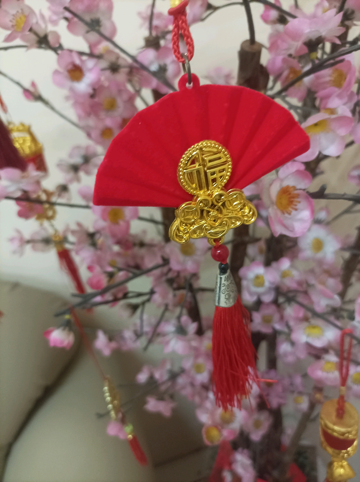 A close-up, slightly blurred shot of a red fan ornament with a gold Chinese character ornament attached, hanging from a decorative branch. The branch is adorned with numerous artificial pink cherry blossoms. Another smaller red tassel ornament is visible to the left, and a golden bell ornament hangs to the right. The background consists of more blurred pink blossoms and a light-colored surface, possibly a wall or furniture. The scene appears to be indoors, decorated for a festive occasion, possibly Chinese New Year, given the traditional ornaments. There are no people or animals present. The lighting is soft and diffused. The location is identified as Bekasi, Indonesia.
