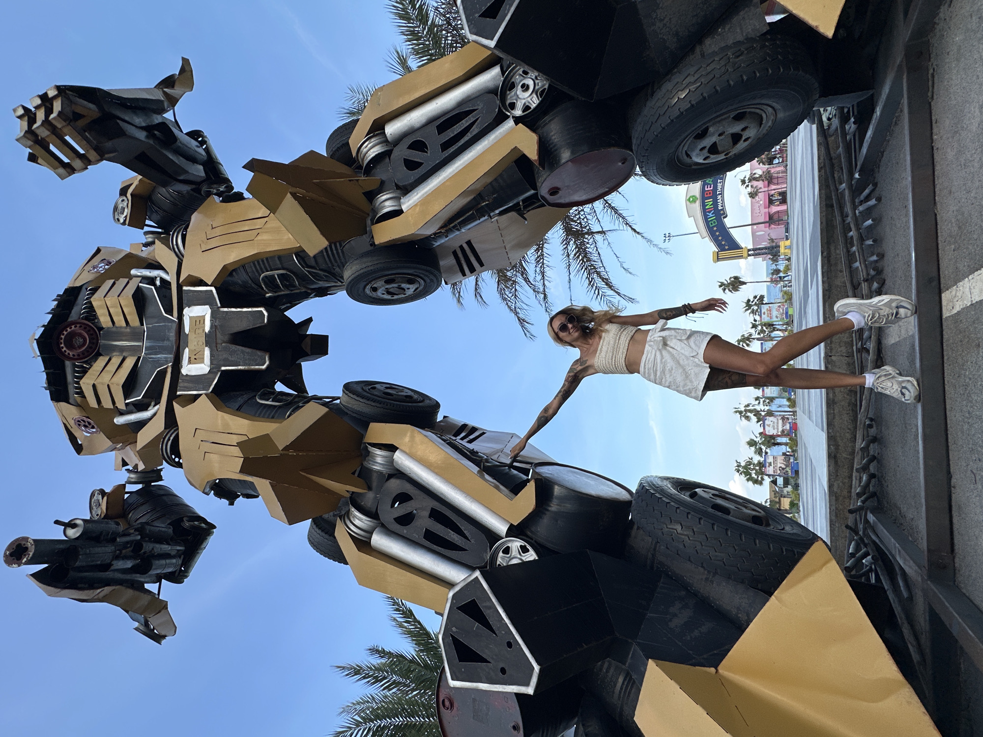 A woman with blonde hair and sunglasses stands beneath a large, golden robot statue in Tien Thanh Ward, Vietnam. She is wearing a white crop top and a white skirt, and she has tattoos on her arms. The robot is made of metal and has large wheels. In the background, there is a sign that says "BIKINI BEACH" and some palm trees. The sky is blue and there are some clouds.