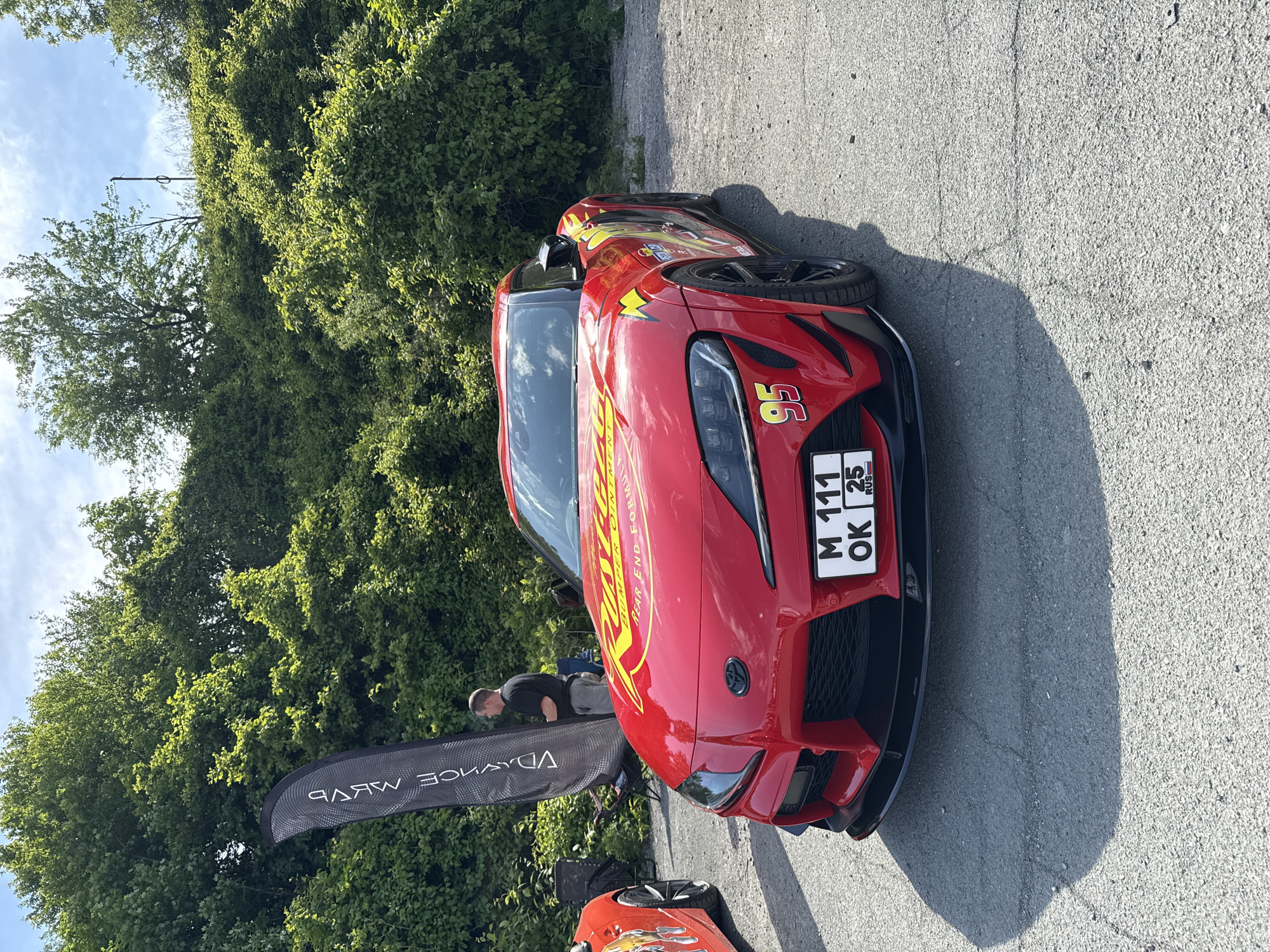 The image shows a red 2020-2023 Toyota Supra customized to resemble Lightning McQueen from the "Cars" franchise, parked on a paved surface adjacent to lush greenery. The car has custom yellow decals and the number "95" on the front. Its license plate reads "M 111 OK 25 RUS". A black teardrop banner with white text, seemingly related to automotive tuning, is visible to the left of the car. A man with short hair, wearing a dark shirt, is standing near the banner. Part of another car is visible behind the Supra. The license plate indicates the vehicle is registered in Vladivostok, Russia.