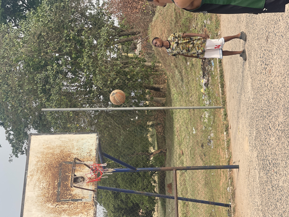 The image depicts an outdoor basketball court in Umunekwu, Nigeria. In the foreground, an asphalt court surface transitions to a chain-link fence and a grassy, unkempt area with scattered litter. Behind this, dense green foliage and trees are visible, with some drier, brown vegetation on the right.
A basketball hoop with a heavily rusted white backboard, a blue metal support frame, and a tattered red and white net stands on the left. To its right, an orange, worn basketball is suspended mid-air.
Two individuals are present. A young Black male, of lean build, stands centrally, facing slightly right and looking upwards towards the basketball. He wears a short-sleeved camouflage-patterned shirt (greens, yellows, browns), white basketball shorts with the number "14" in red on the right leg, and dark slide sandals. A second individual, partially visible on the far right, is a Black male wearing a black and green sleeveless jersey; only his right shoulder, arm, and part of his back are within the frame. A thin, grey metal pole stands vertically behind the chain-link fence, to the right of the basketball hoop.